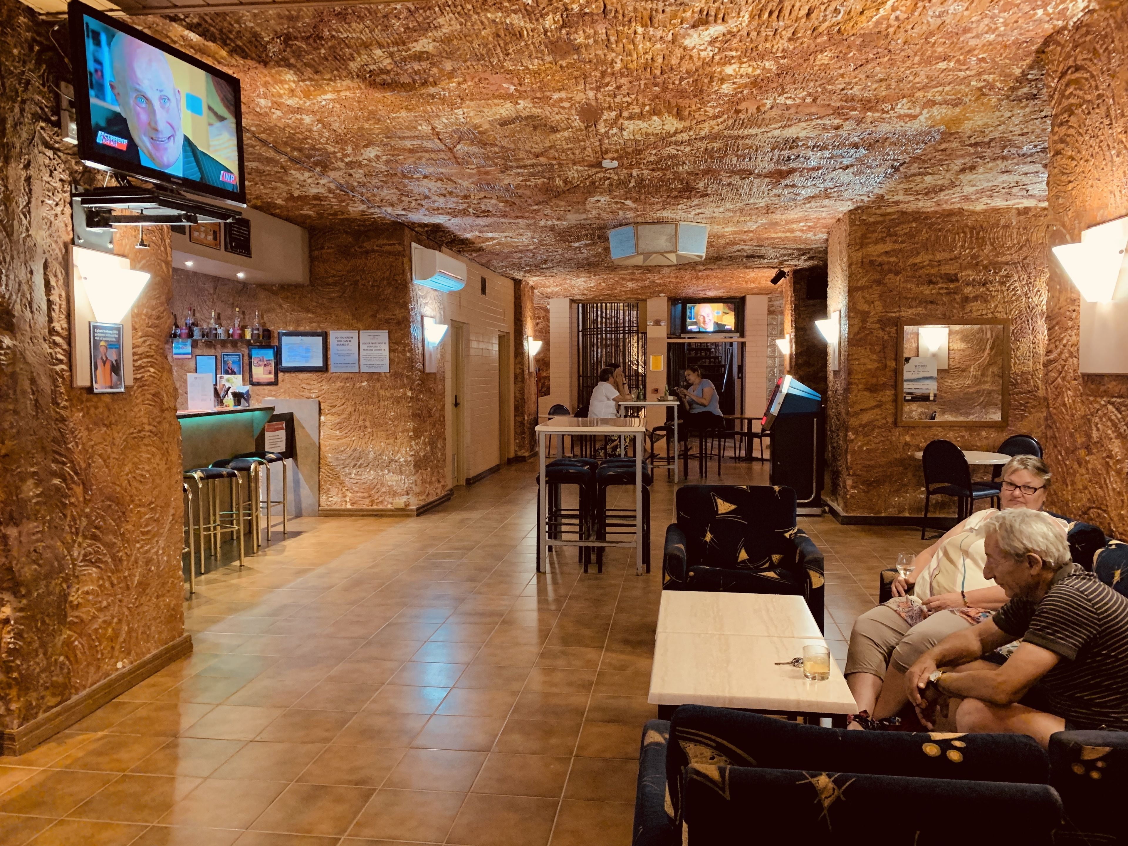 Een restaurant met een rustieke inrichting, zichtbaar aan de stenen muren en het houten plafond. Er zijn tafels en stoelen voor de gasten, en een bar met een toonbank en flessenkasten.