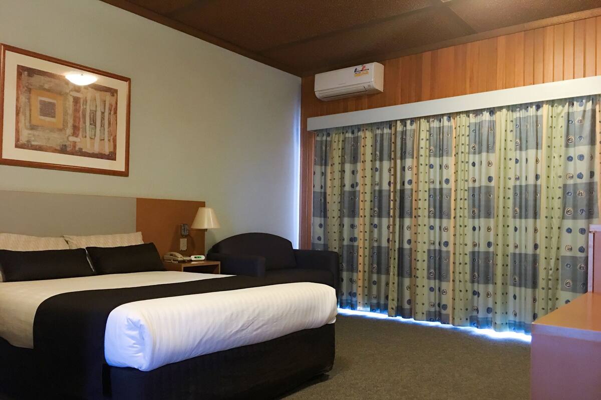 A hotel room with a large bed, a nightstand, a lamp, a chair, a window with curtains, and an air conditioning unit.