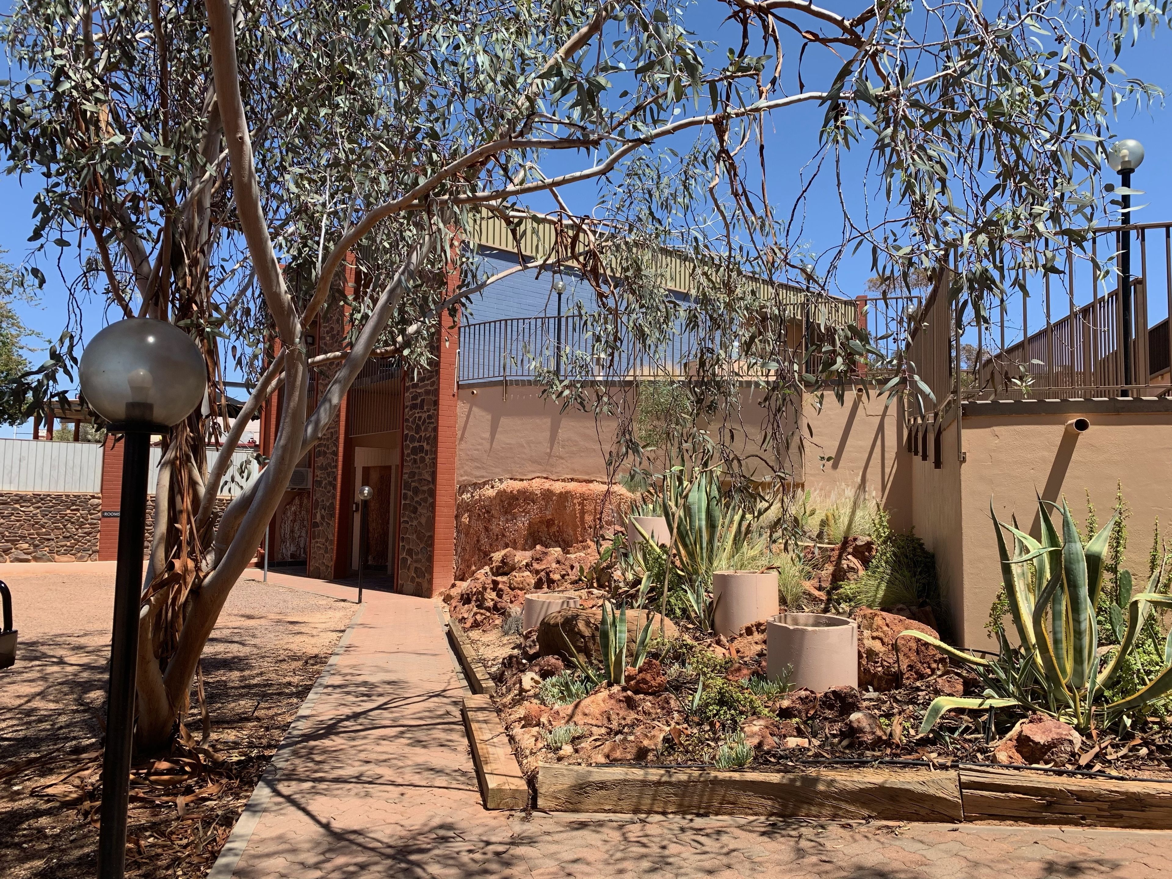 A pathway leading to a building with a stone facade, surrounded by desert plants and a metal fence.