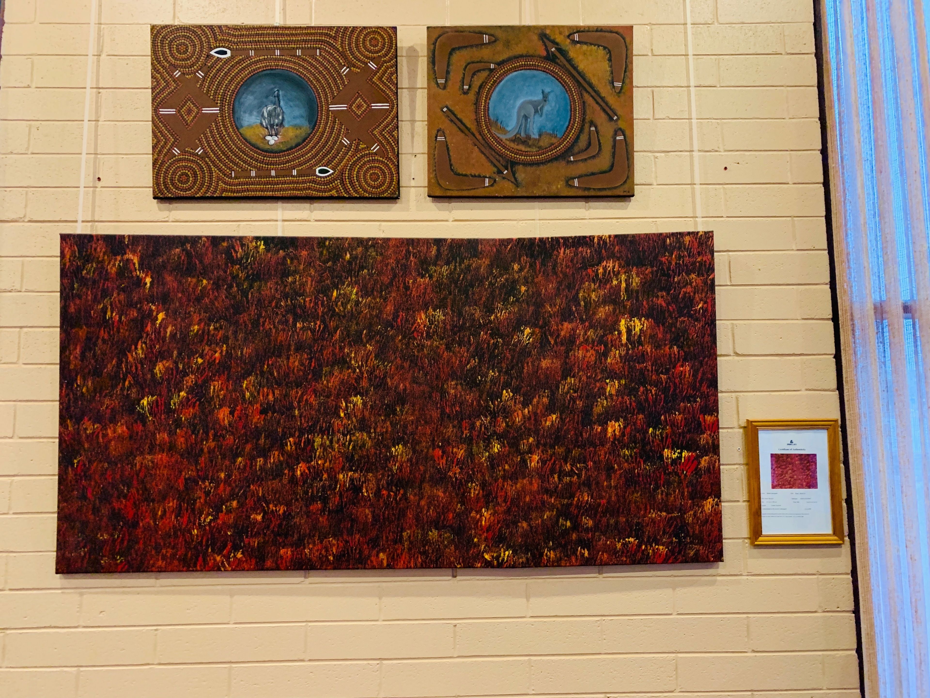 Een muur met drie kunstwerken: twee ingelijste schilderijen met Aboriginal designs en een groot abstract schilderij.