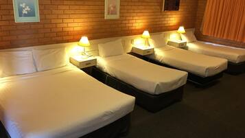 A hotel room with four single beds, each with a bedside table and lamp. The room has a brick wall with framed artwork above the beds.