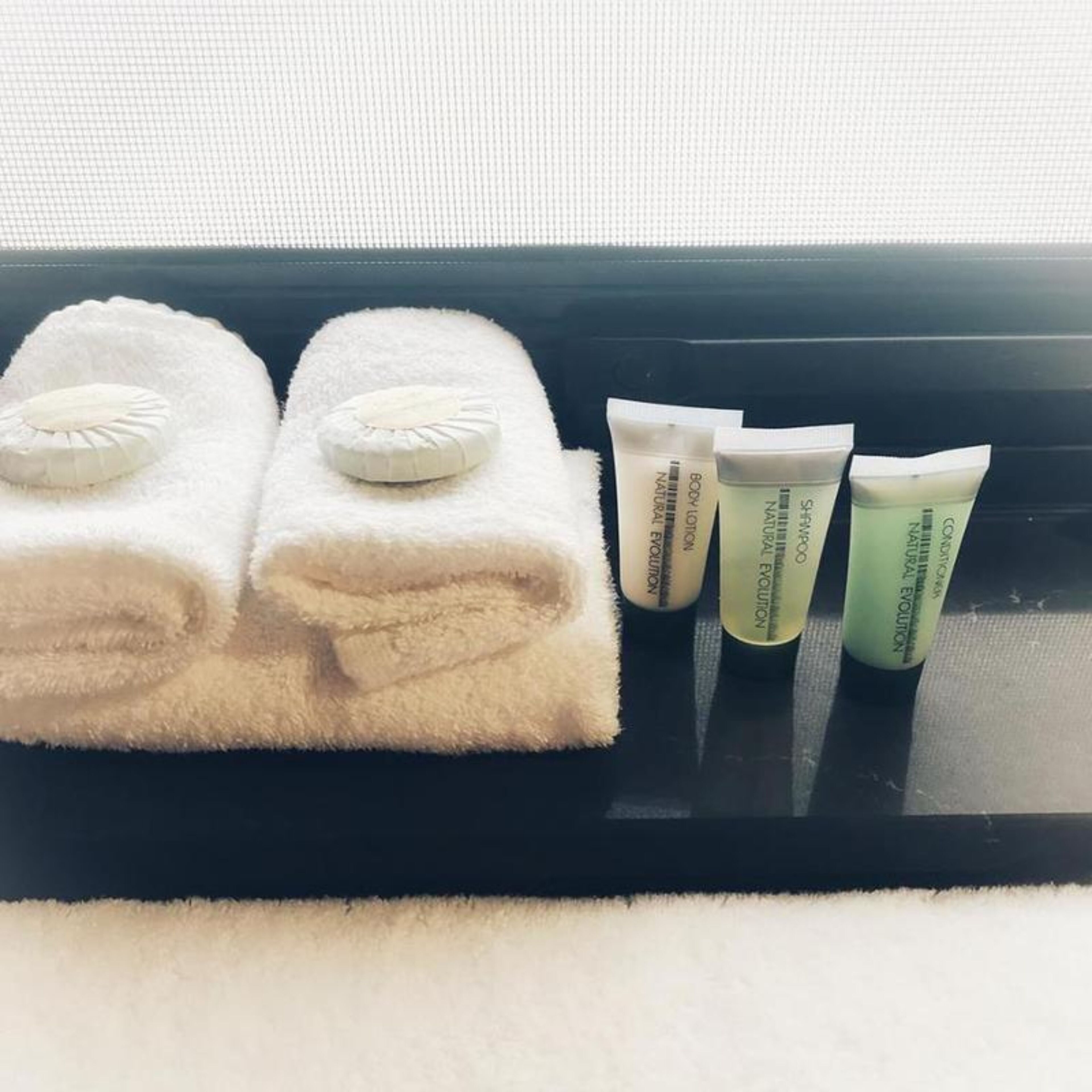 Un plateau noir avec deux serviettes blanches et trois tubes de lotion pour le corps, posés sur un rebord de fenêtre.