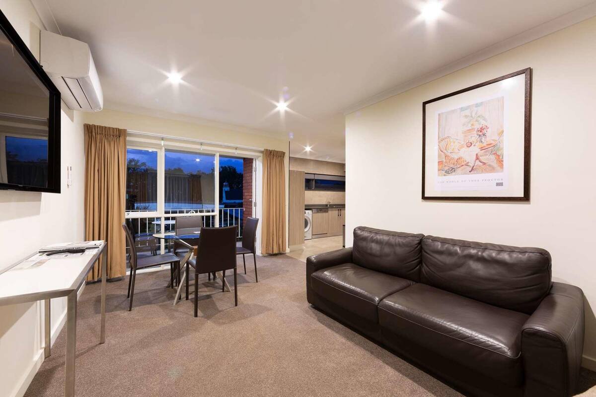 A modern living room with a leather sofa, a dining table with chairs, a flat-screen TV mounted on the wall, and a large window with curtains.
