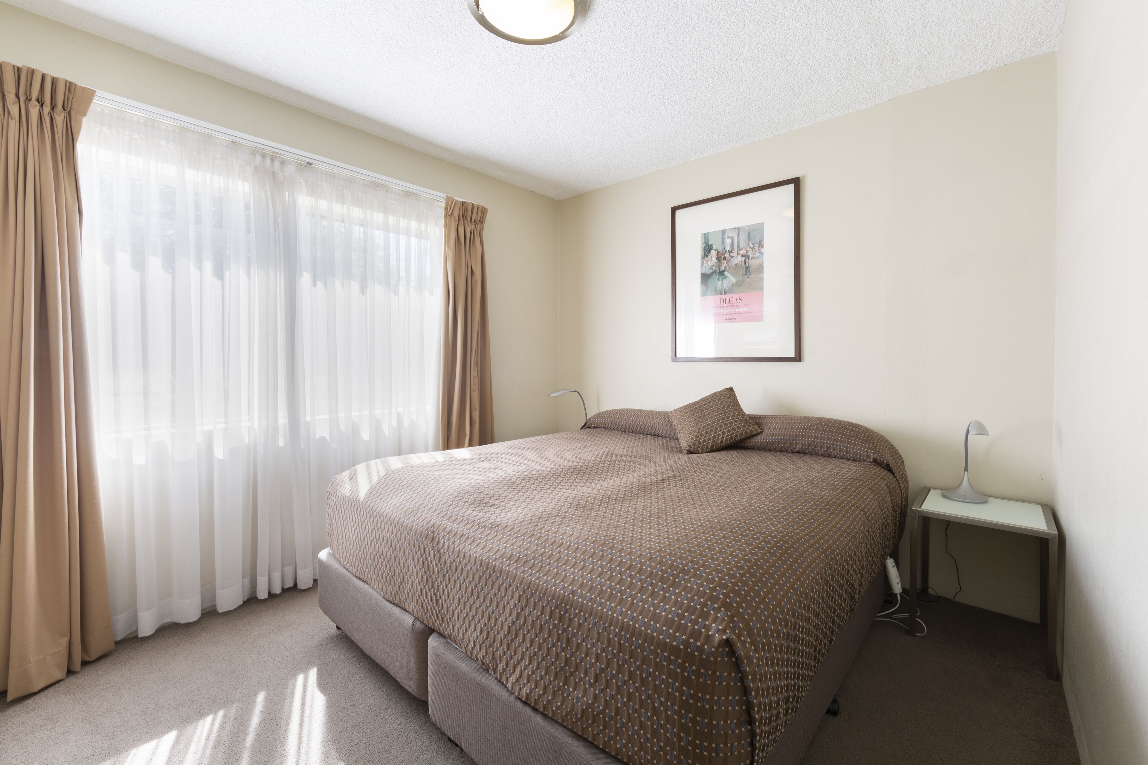 Un dormitorio con cama, cortinas, una mesita de noche con lámpara y un cuadro enmarcado en la pared.