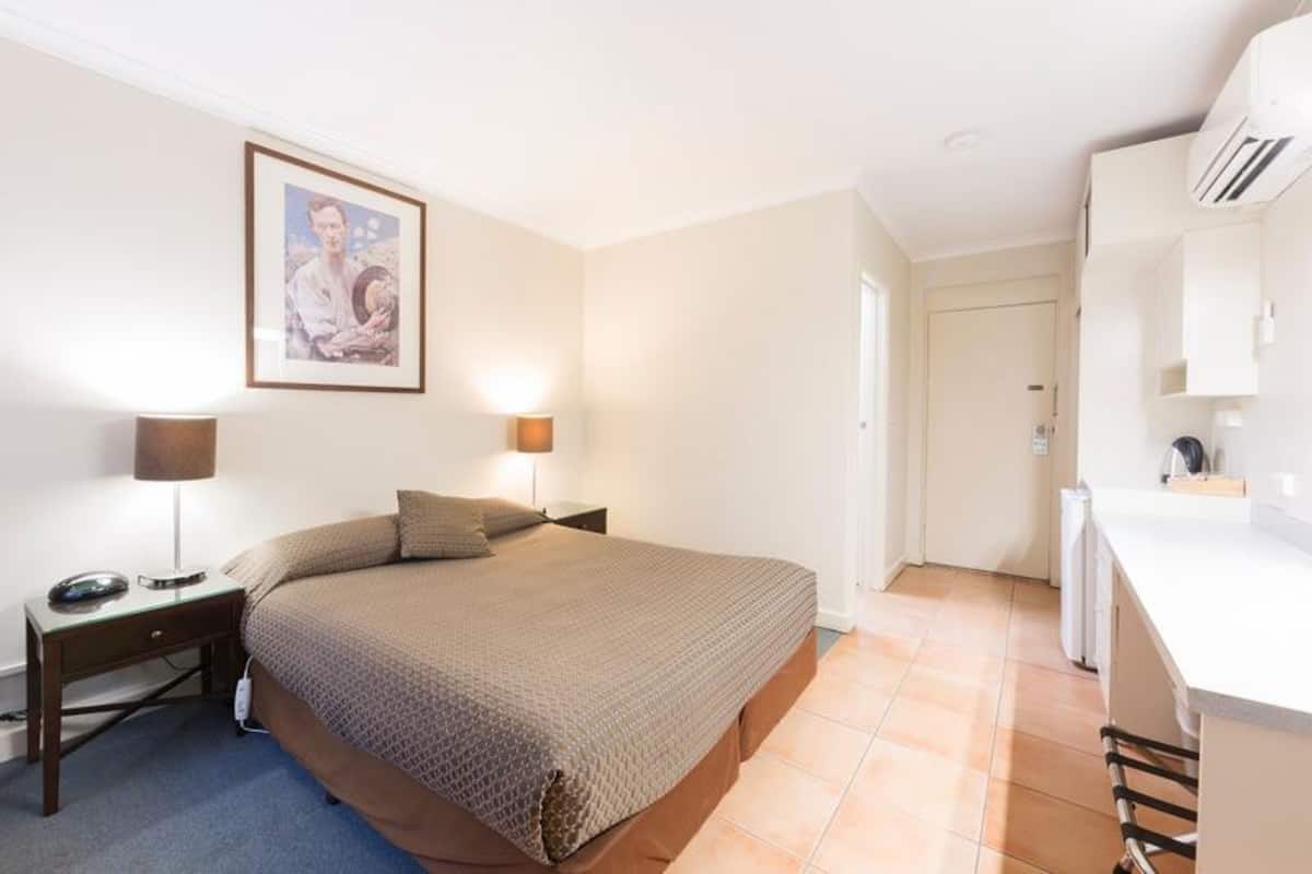 A bedroom with a bed, bedside lamps, a framed picture on the wall, and a kitchenette area.