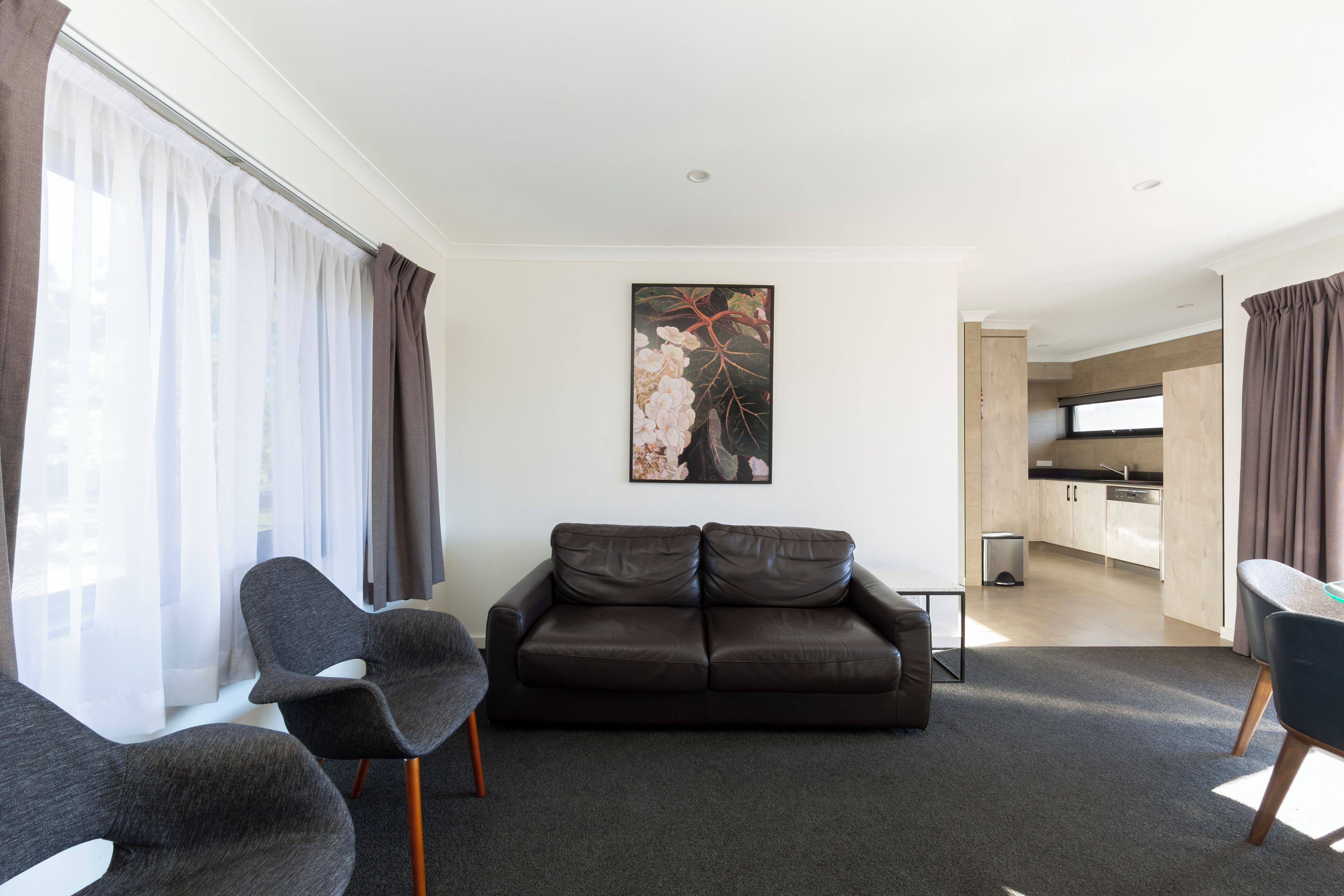 A modern living room with a sofa, chairs, a painting, and a view into the kitchen.