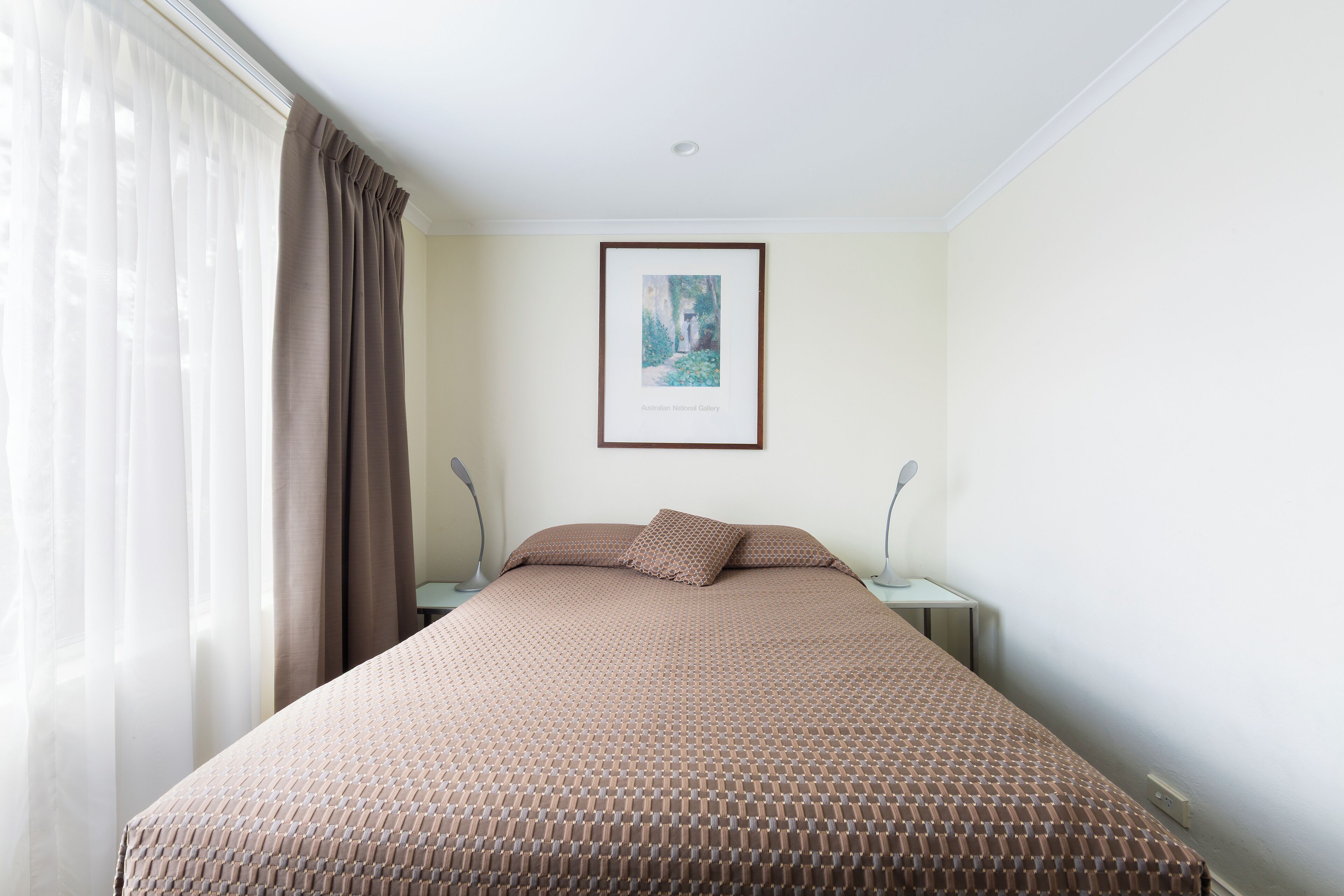 Quarto com cama, duas mesas de cabeceira, um quadro na parede e janela com cortinas.