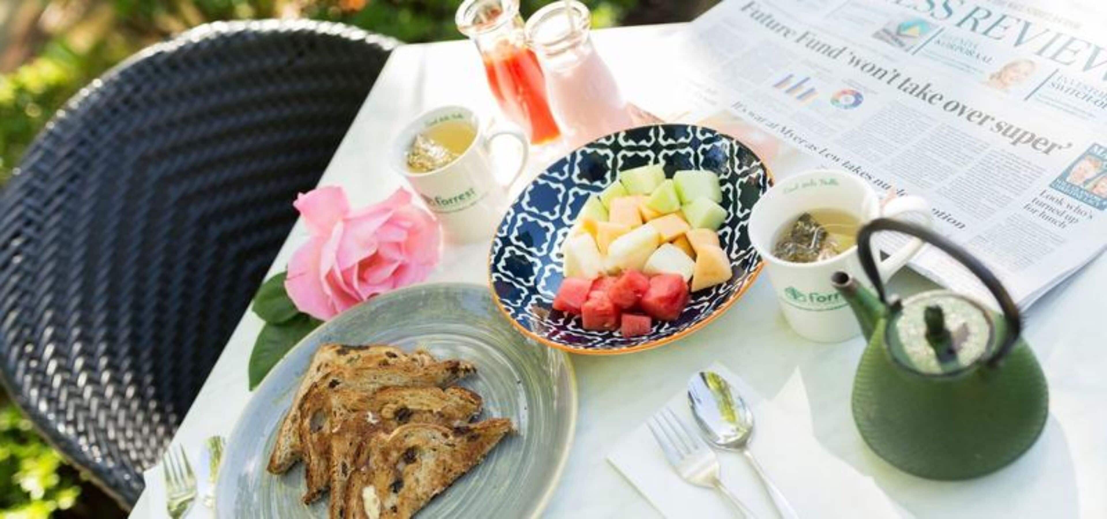 A table with a newspaper, a teapot, a plate with toast, a bowl of fruit, and a pink flower.