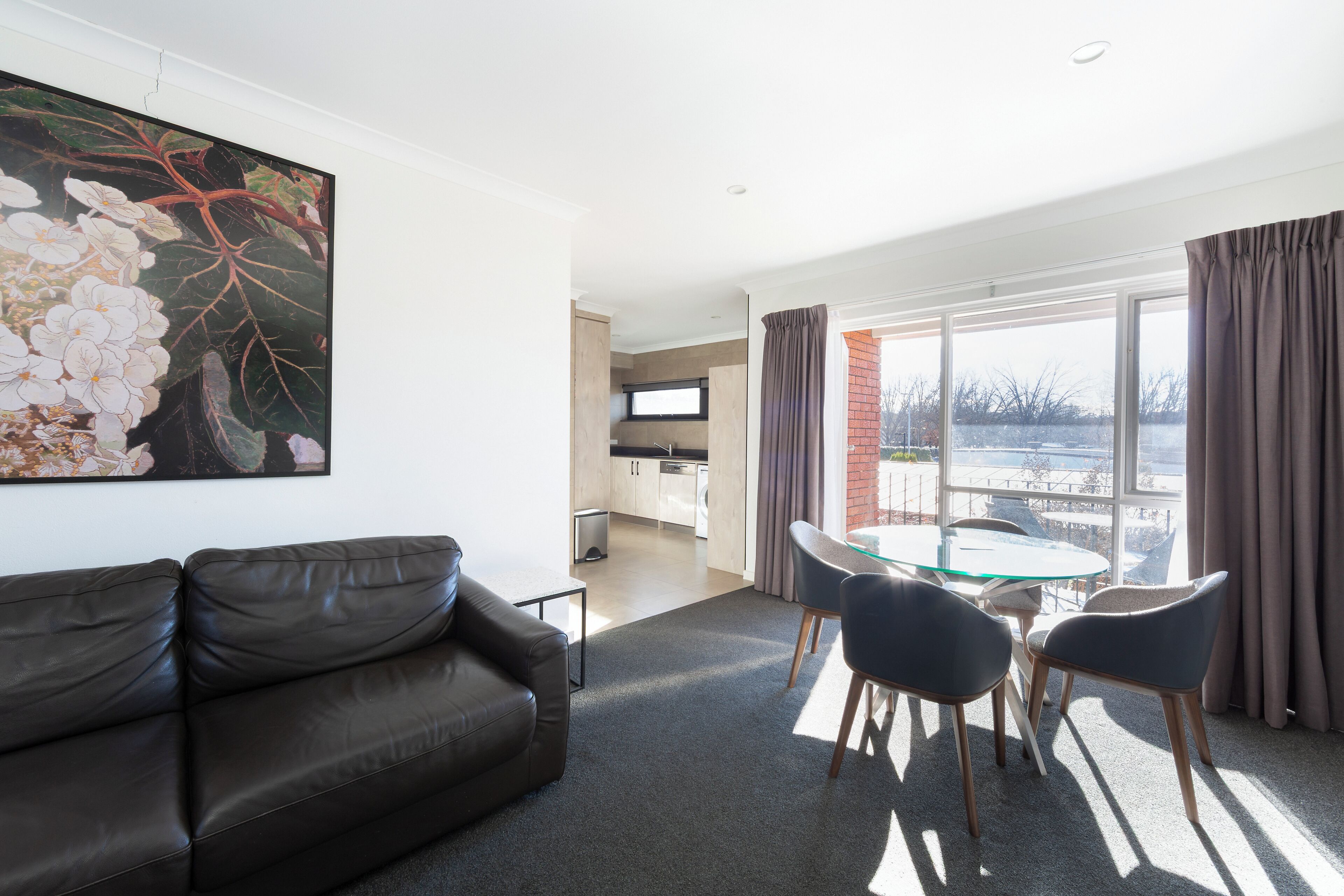 A modern living room with a leather sofa, a glass table with chairs, and a large window with curtains.