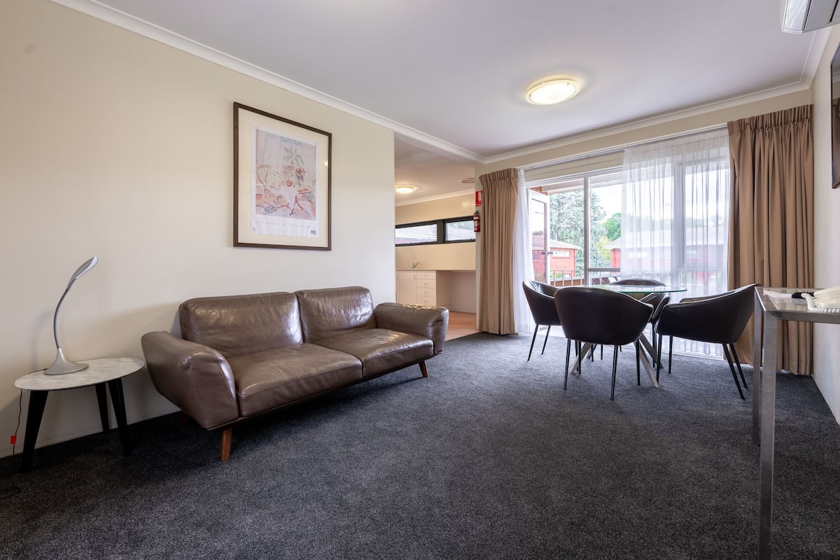 A modern living room with a leather sofa, a small side table, a dining area with a table and chairs, and a large window with curtains.