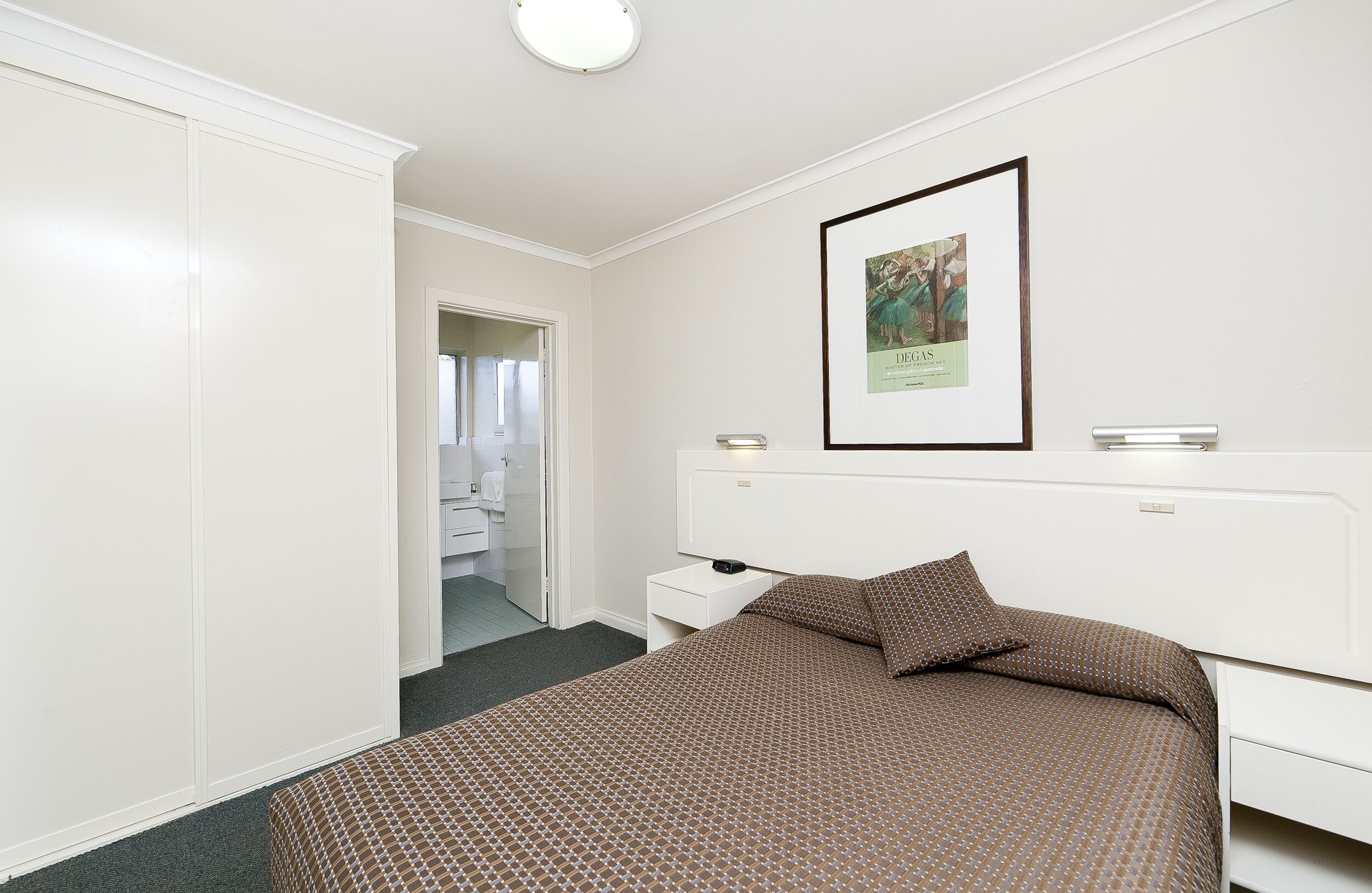 A hotel room with a large bed, a white wardrobe, a framed picture on the wall, and a bedside table with a lamp.