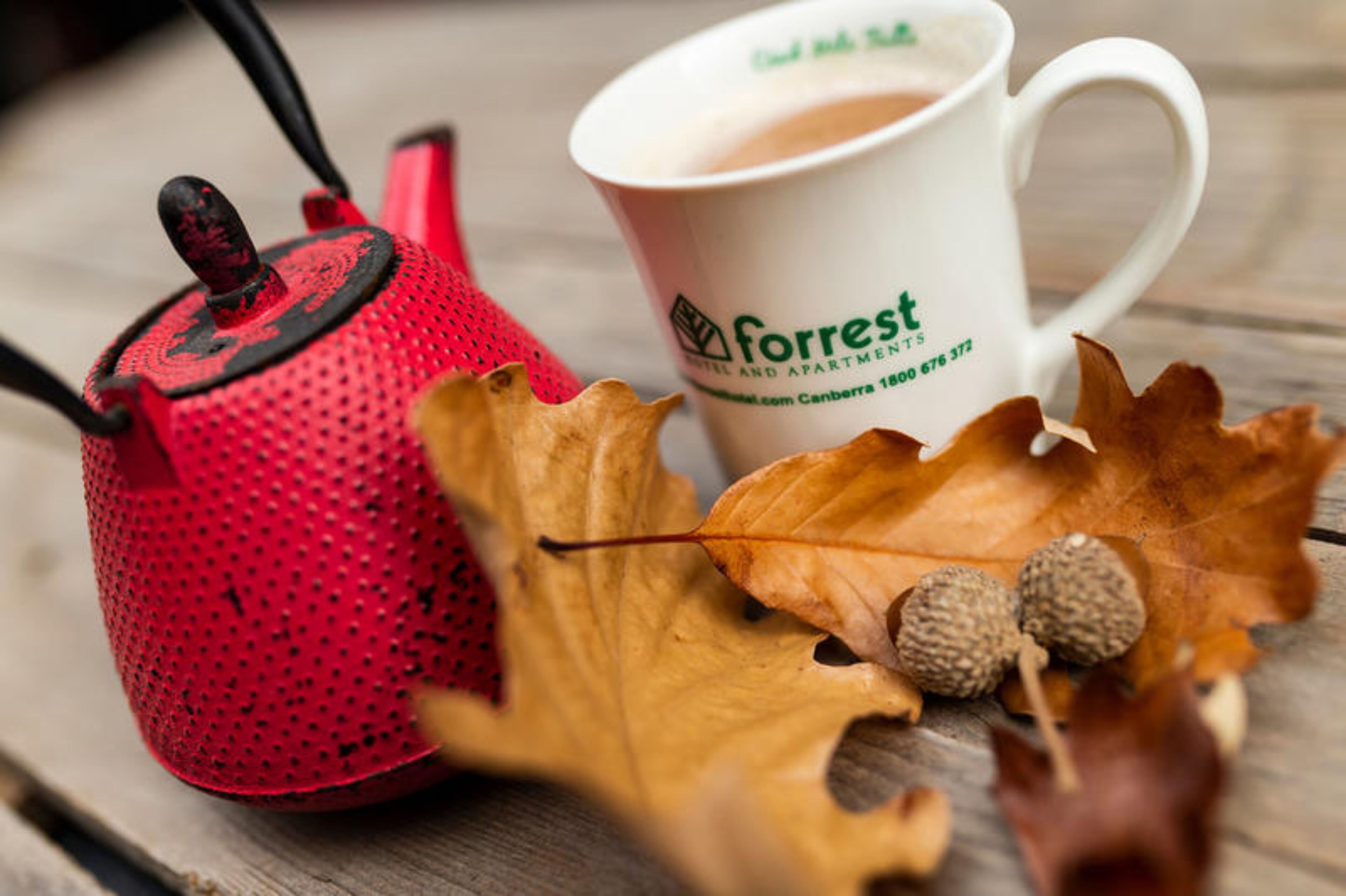 Una tetera roja, una taza blanca con la marca 'Forrest' y hojas otoñales sobre una superficie de madera.