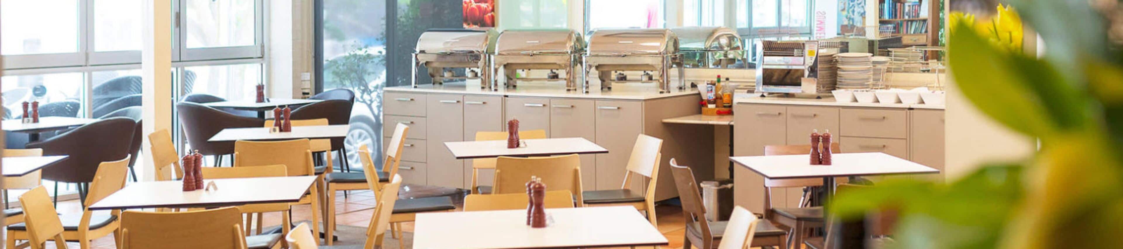A restaurant interior with multiple tables and chairs, a kitchen area with appliances, and a view through large windows.