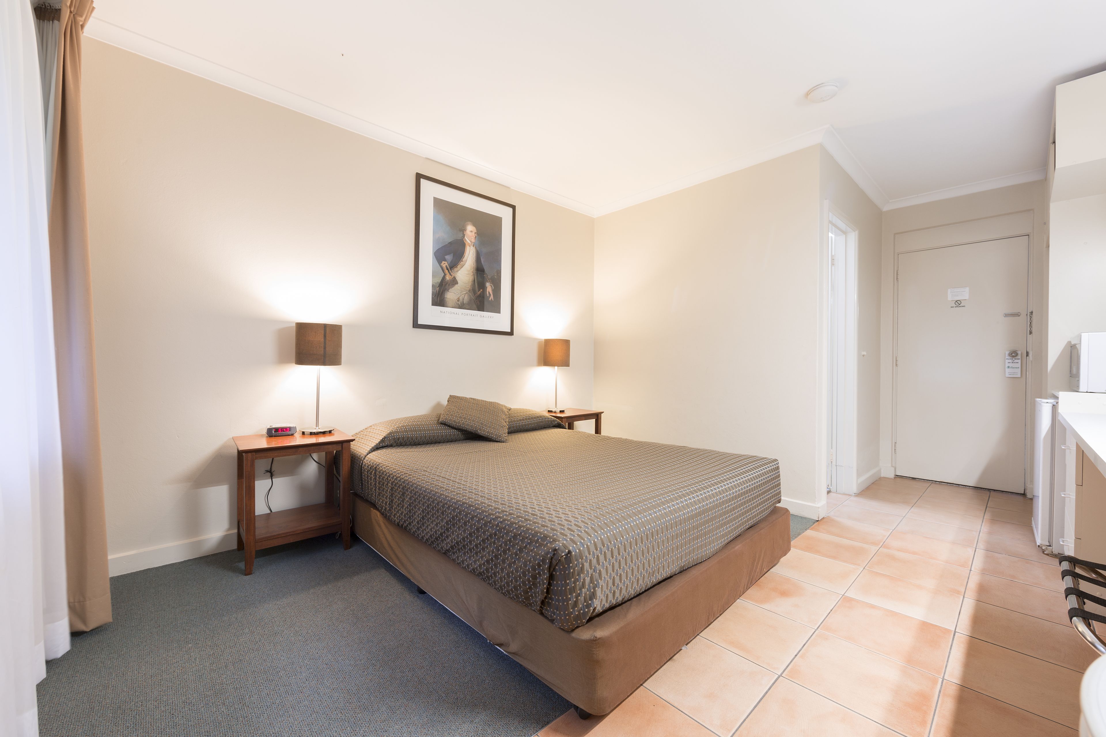A bedroom with a bed, bedside tables, a lamp, a framed picture on the wall, and a door leading to another room.