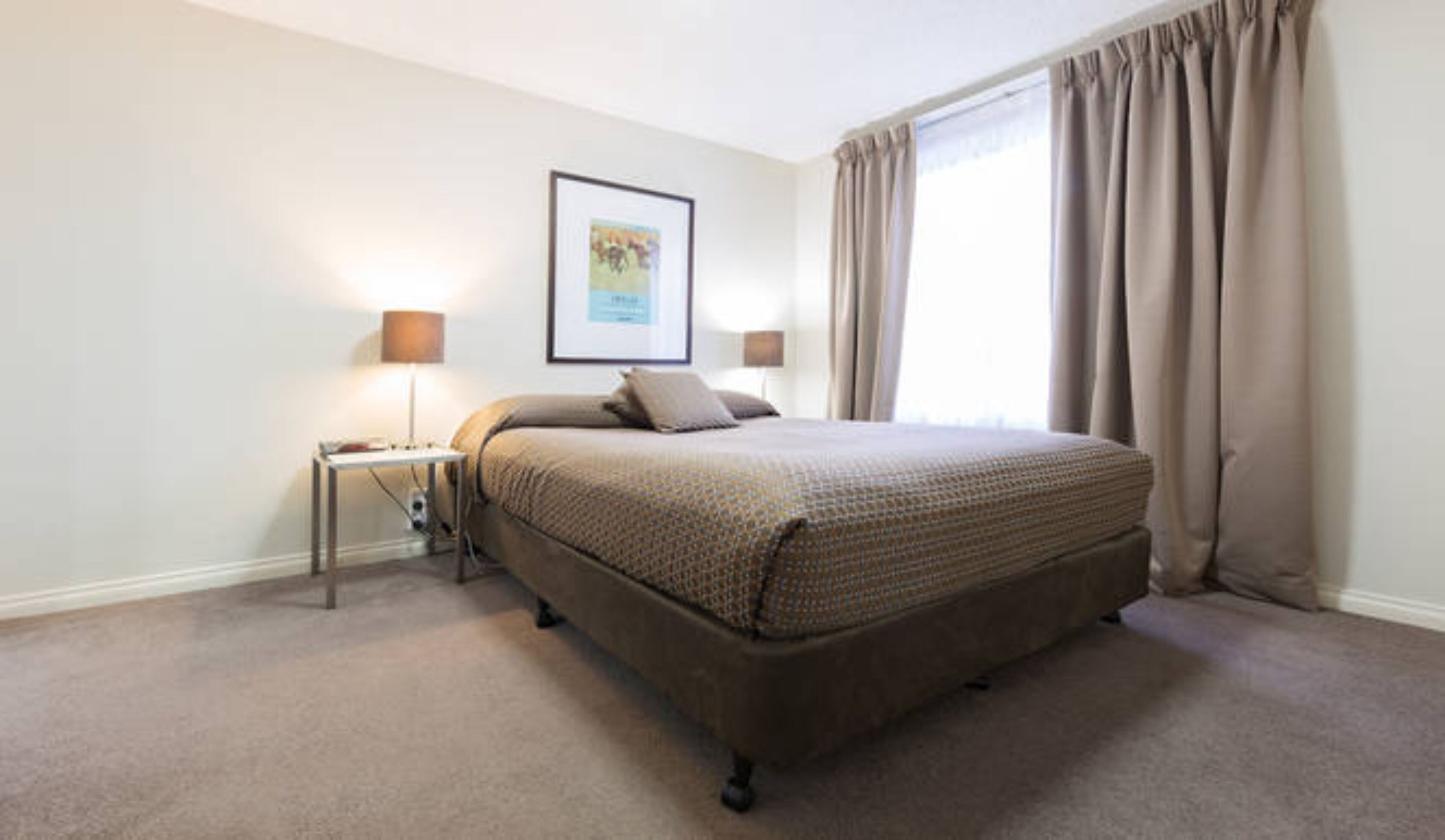 A bedroom with a large bed, two bedside tables with lamps, a framed picture on the wall, and a window with curtains.