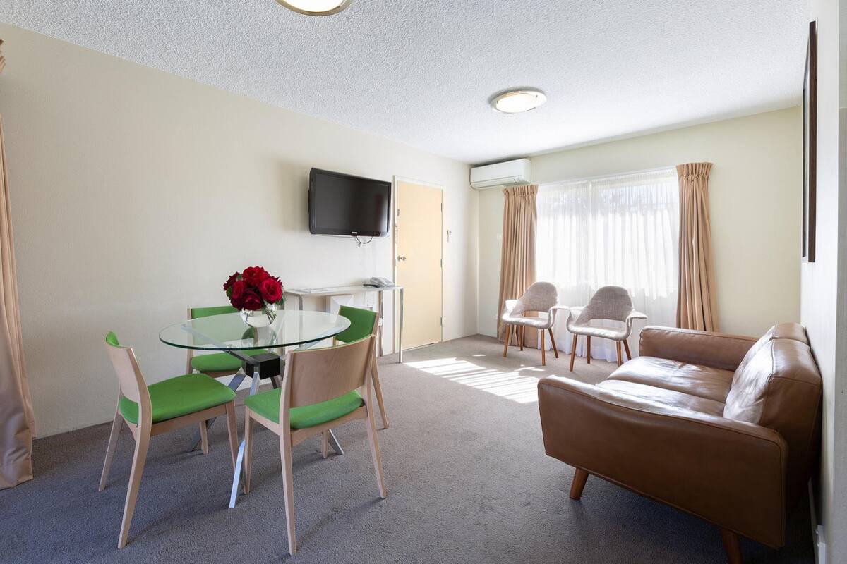 A modern living room with a glass table, green chairs, a brown sofa, and a flat-screen TV mounted on the wall.
