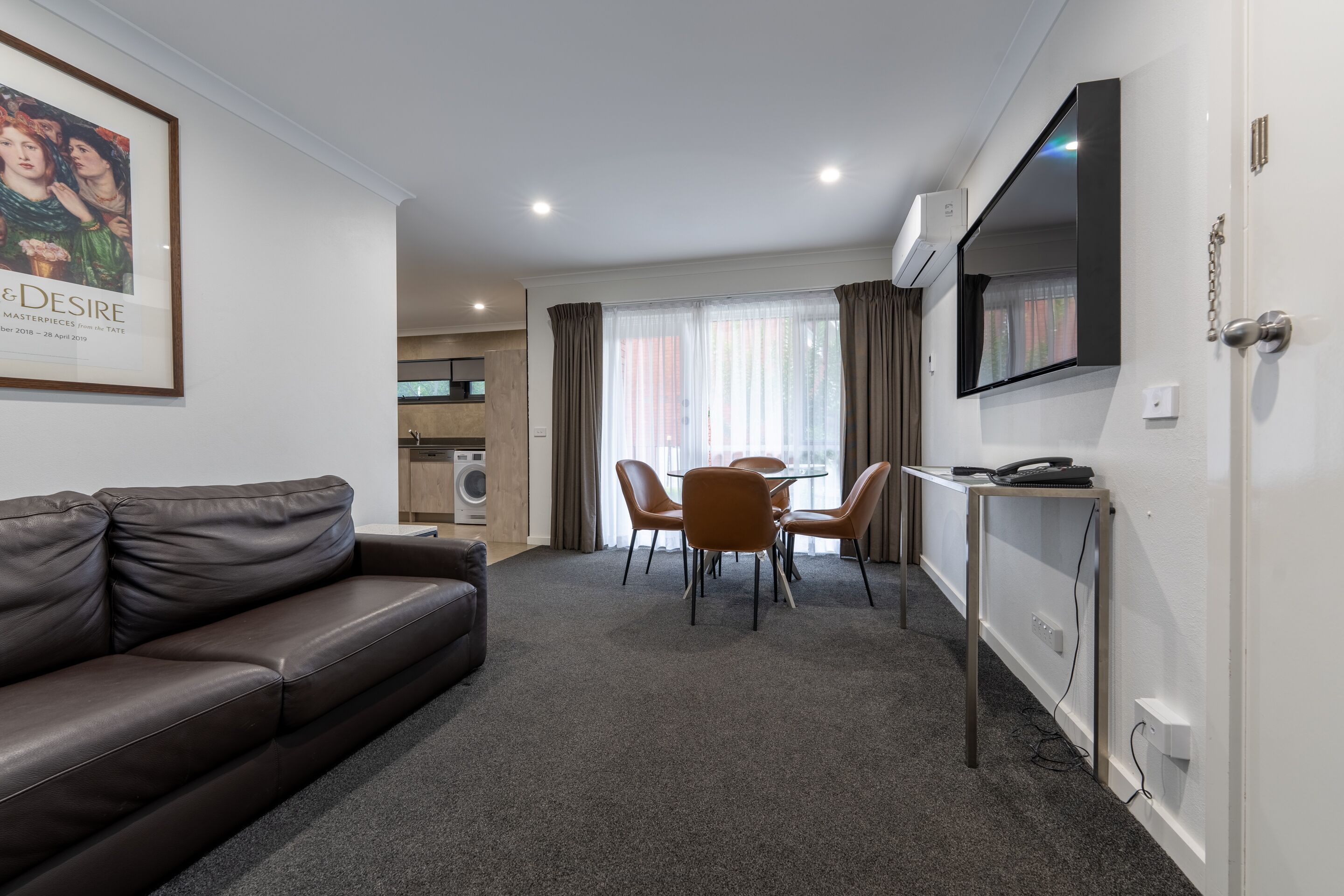 A modern living room with a brown leather sofa, a dining table with four chairs, a TV mounted on the wall, and a kitchen area in the background.