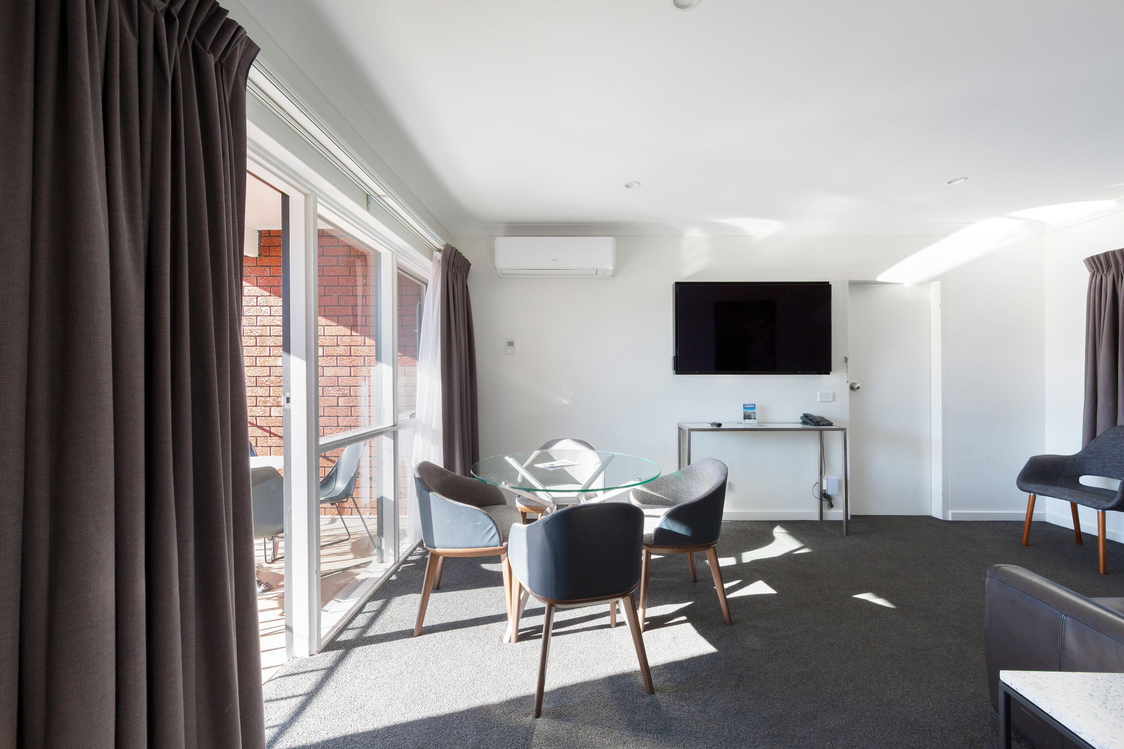A modern room with a glass table, chairs, a flat-screen TV, and a view of a brick building.