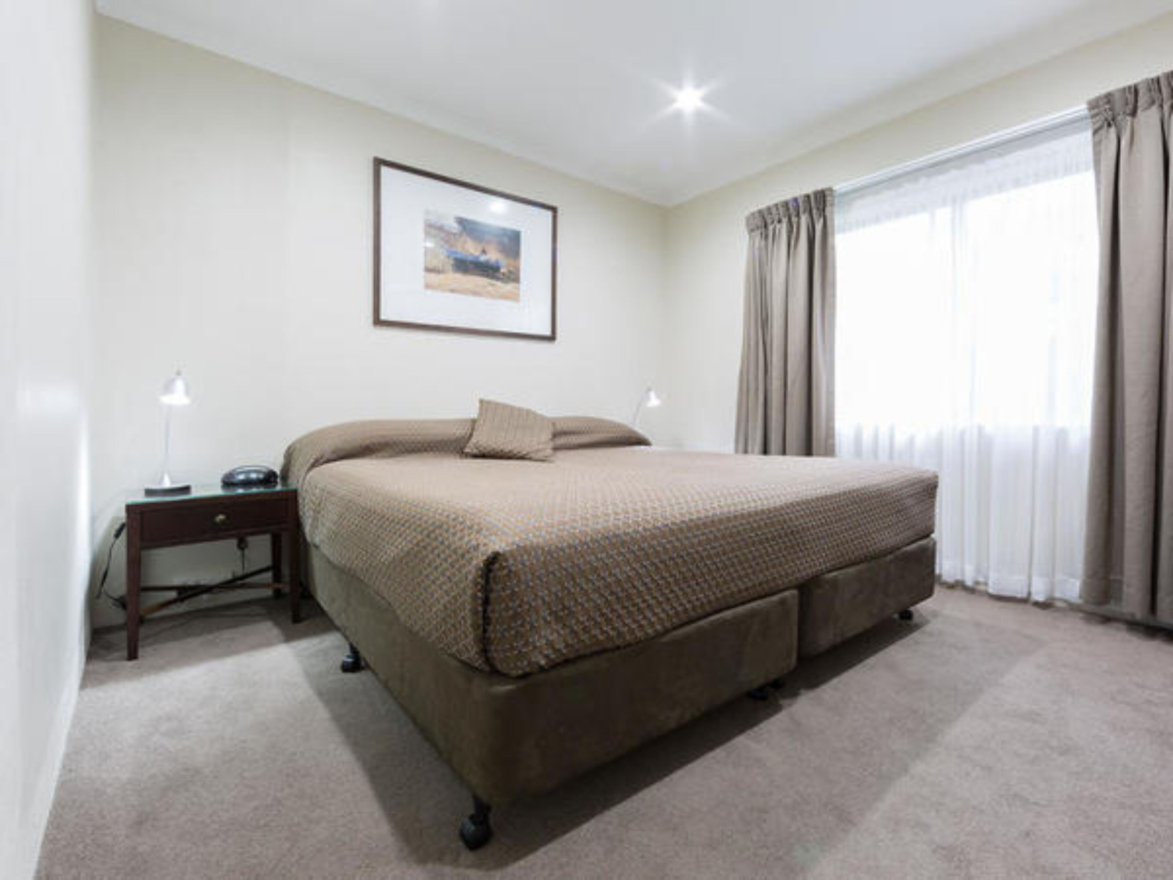 A bedroom with a bed, a bedside table with a lamp, a framed picture on the wall, and a window with curtains.