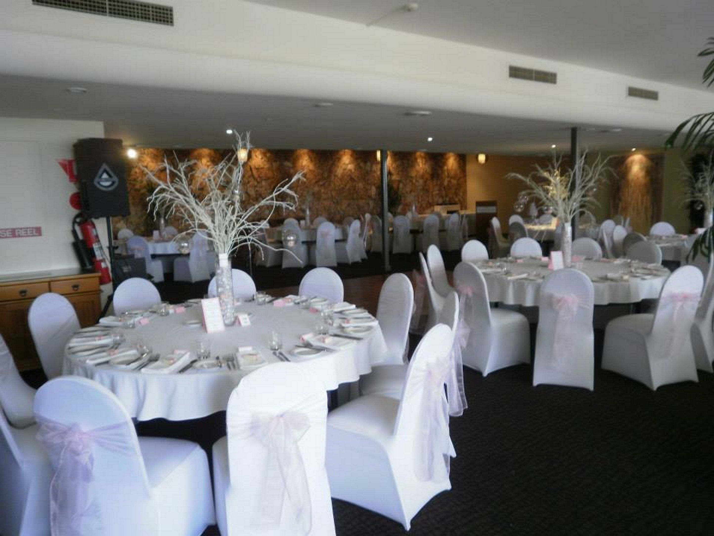 Un salón de banquetes con mesas redondas, sillas blancas adornadas con lazos y ramas blancas decorativas.