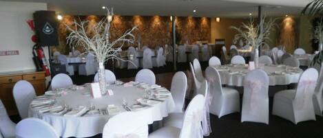 Un salón de banquetes con mesas redondas, sillas blancas adornadas con lazos y ramas blancas decorativas.