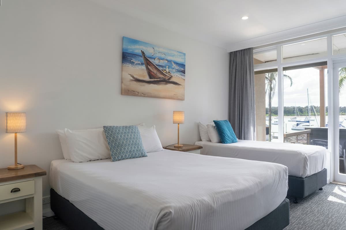 A hotel room with two beds, a painting of a sailboat on the wall, and a view of the beach and boats through the window.