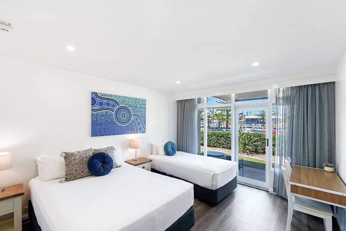 A hotel room with two beds, a desk, a chair, a window with curtains, and a wall art piece.