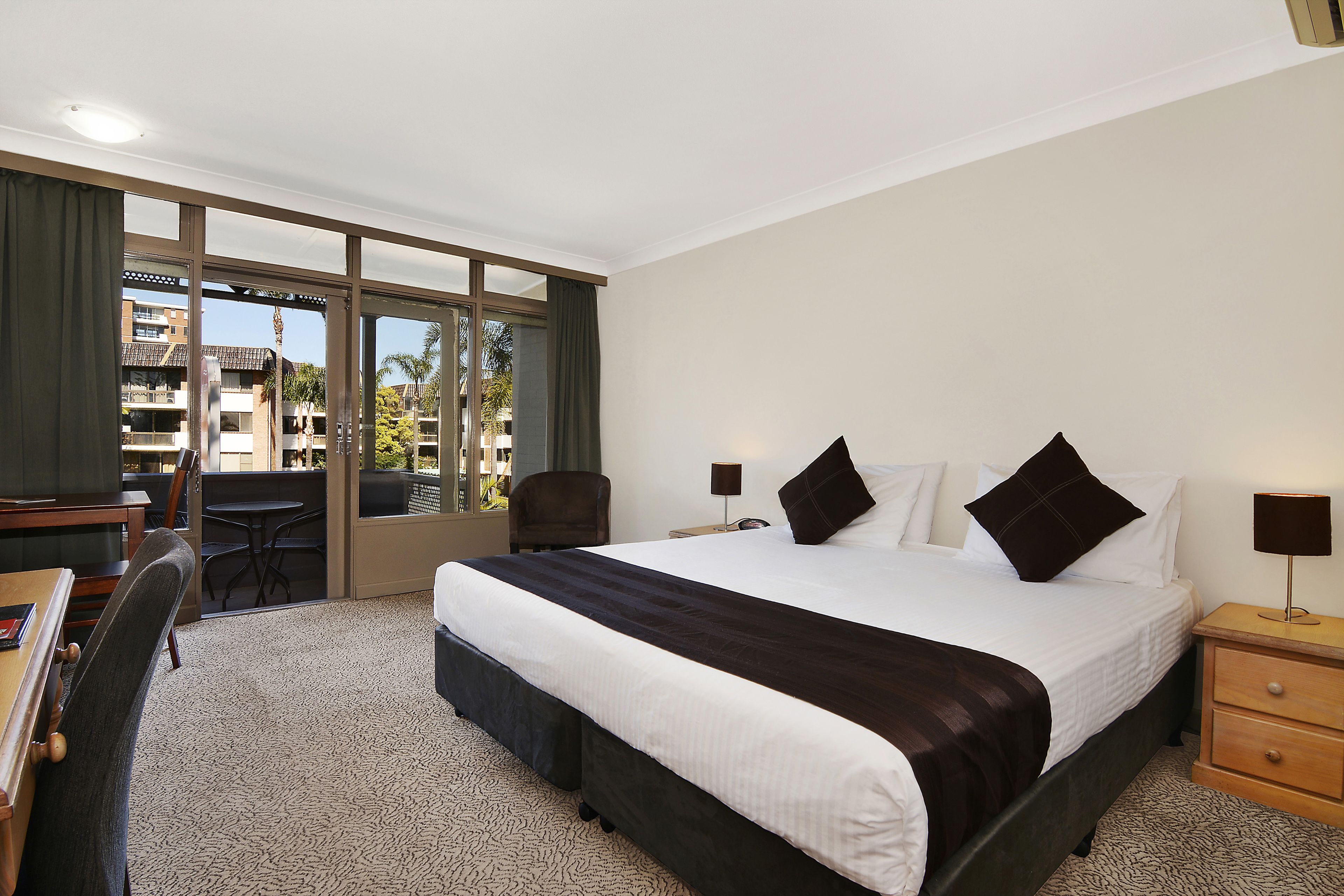 A hotel room with a large bed, a desk, a chair, a nightstand, and a view of the outside.