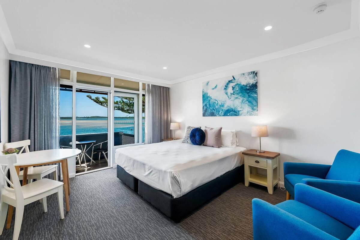 A hotel room with a bed, a desk, a chair, a nightstand, a lamp, and a view of the ocean.