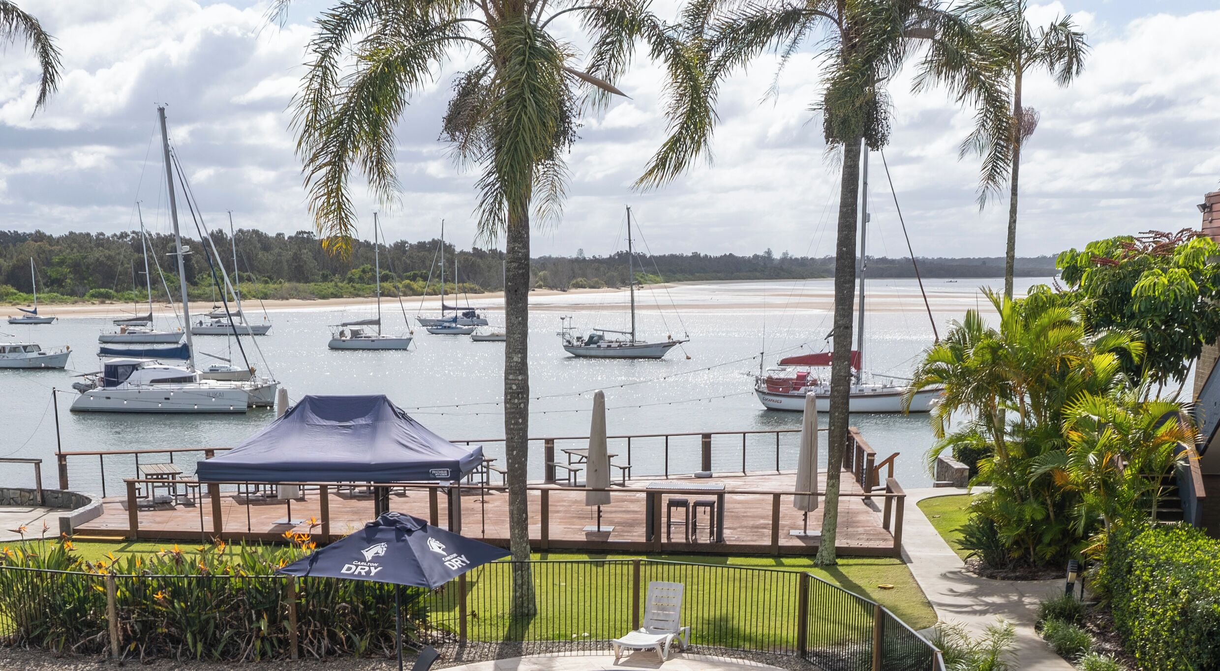 A marina with multiple sailboats docked, a beach in the background, and a covered seating area with a tent.