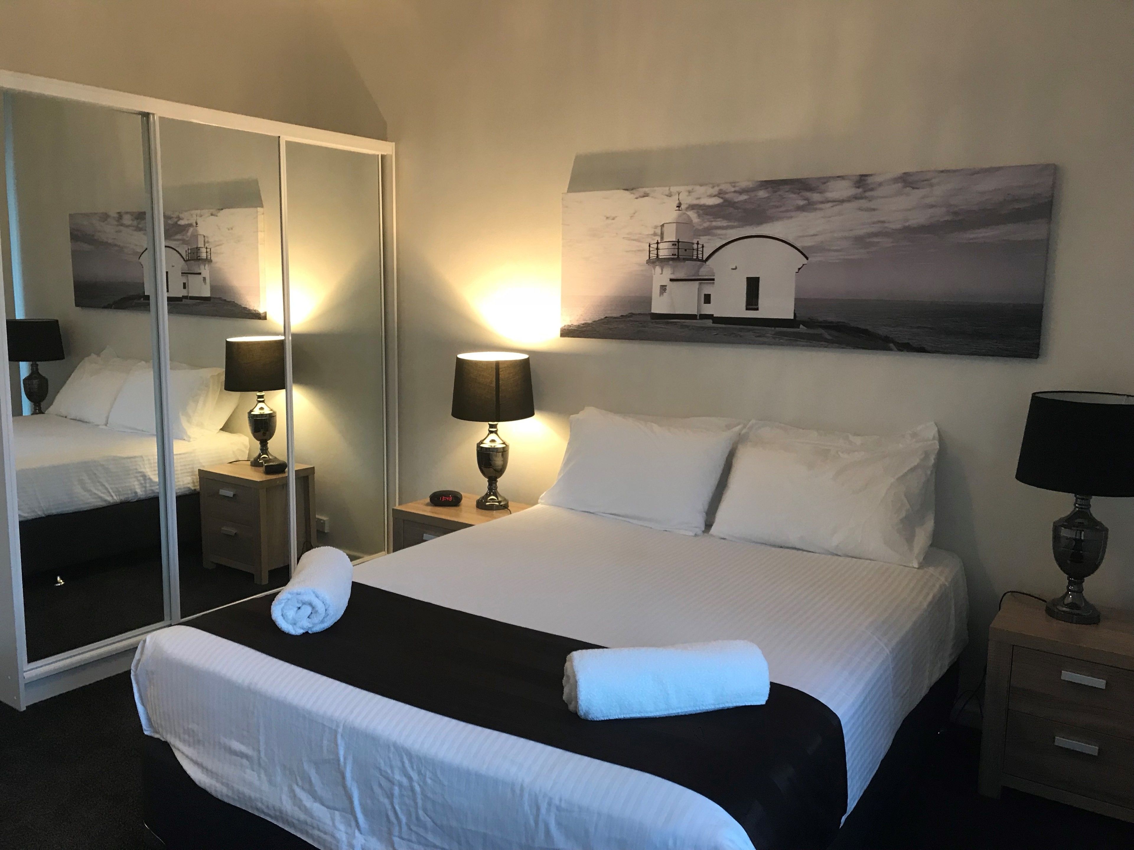Un lit bien fait, avec du linge de lit blanc, deux tables de chevet avec des lampes, un grand miroir et un tableau représentant un paysage côtier.