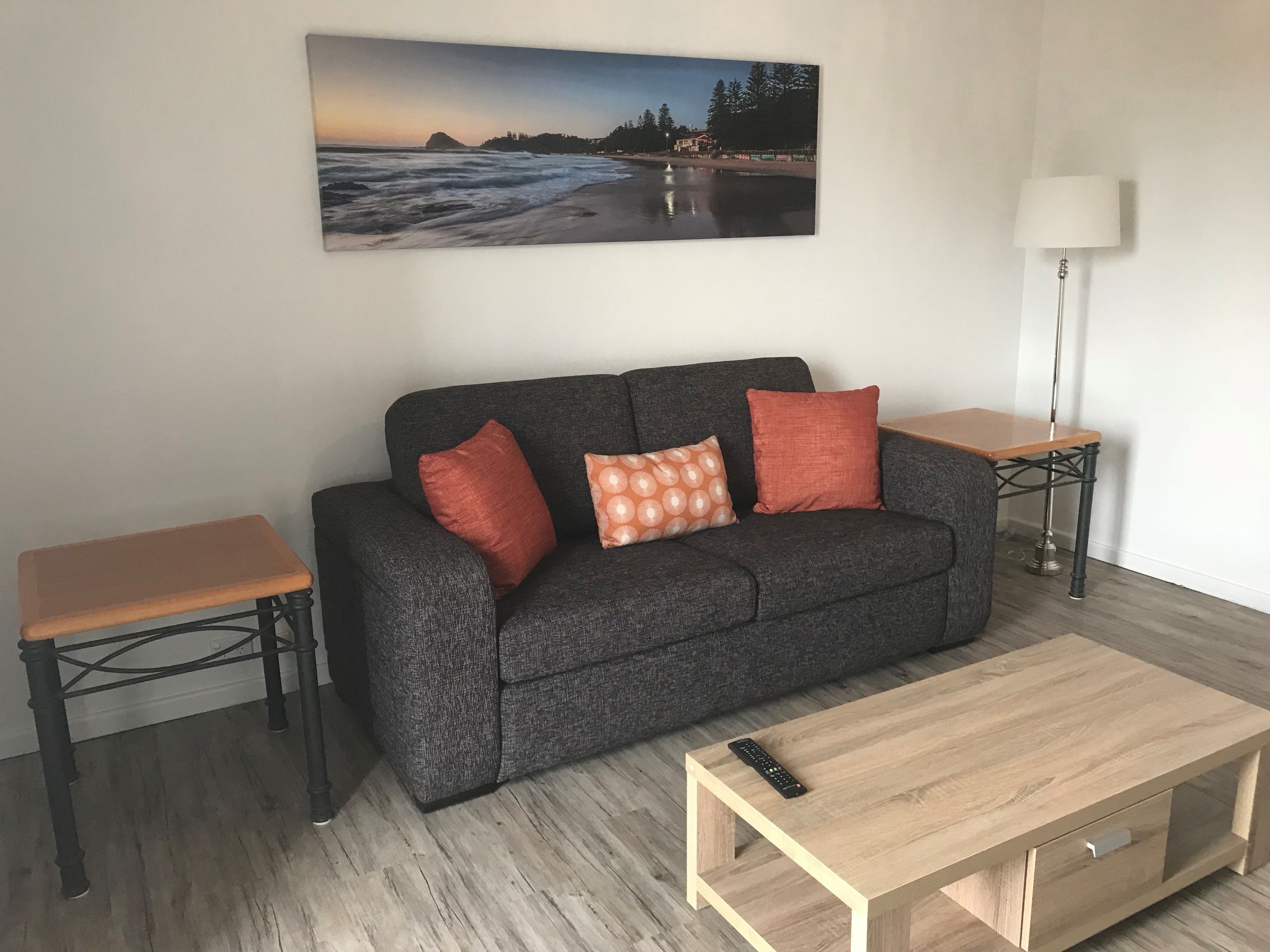 Una sala de estar con un sofá gris, dos mesitas auxiliares, una mesa de centro de madera y un cuadro con una escena de playa en la pared.