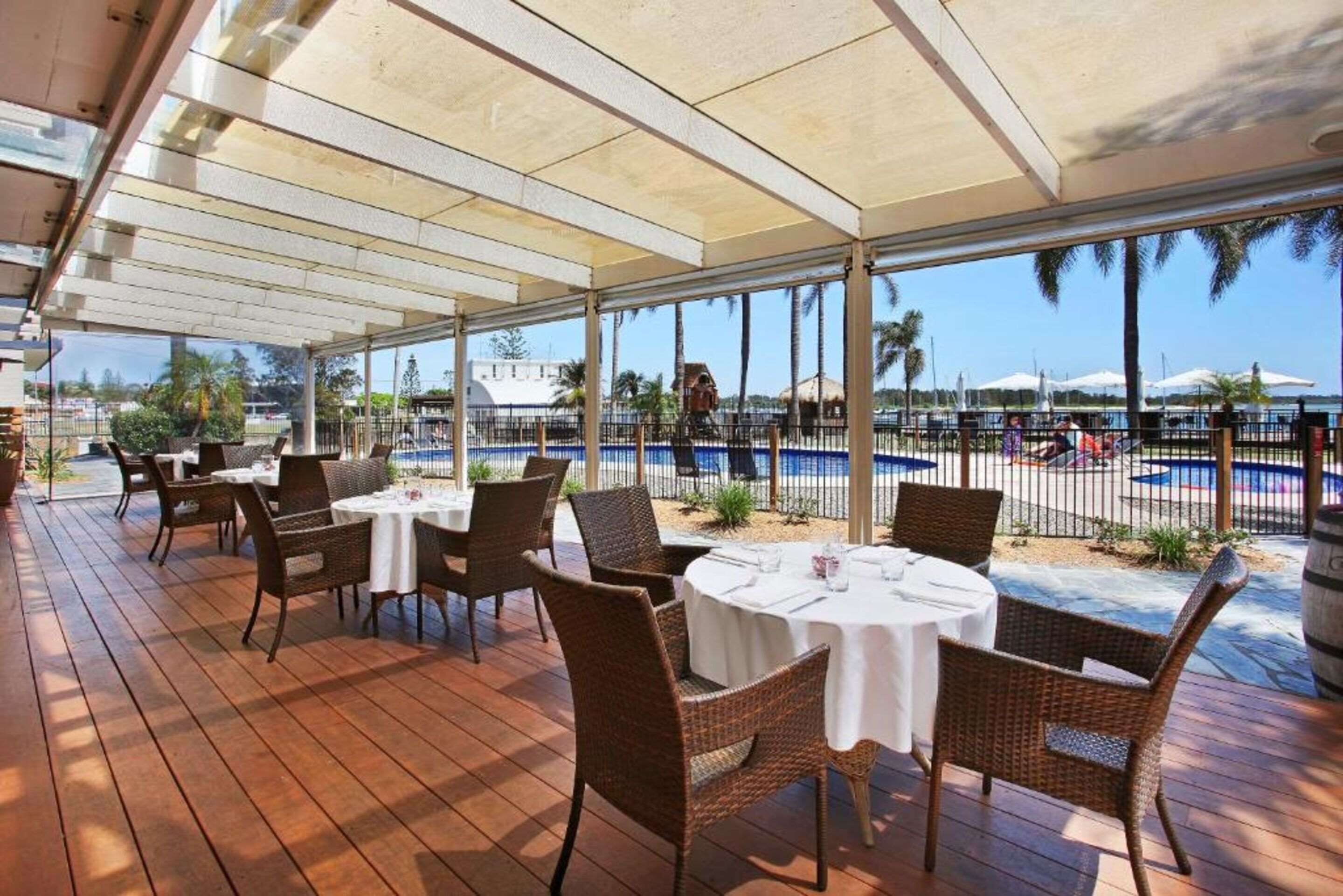 An outdoor dining area with tables and wicker chairs, overlooking a pool and palm trees.