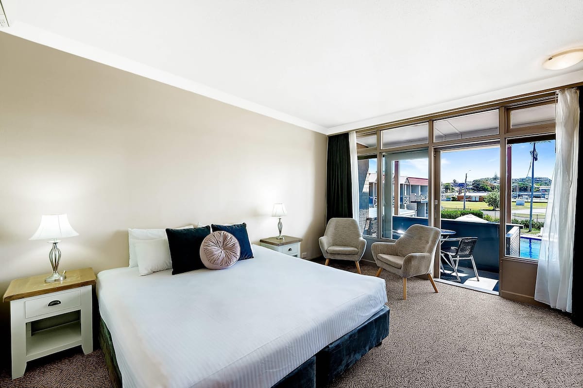 A hotel room with a large bed, two bedside tables with lamps, a chair, a small table, and a view of a swimming pool and buildings.