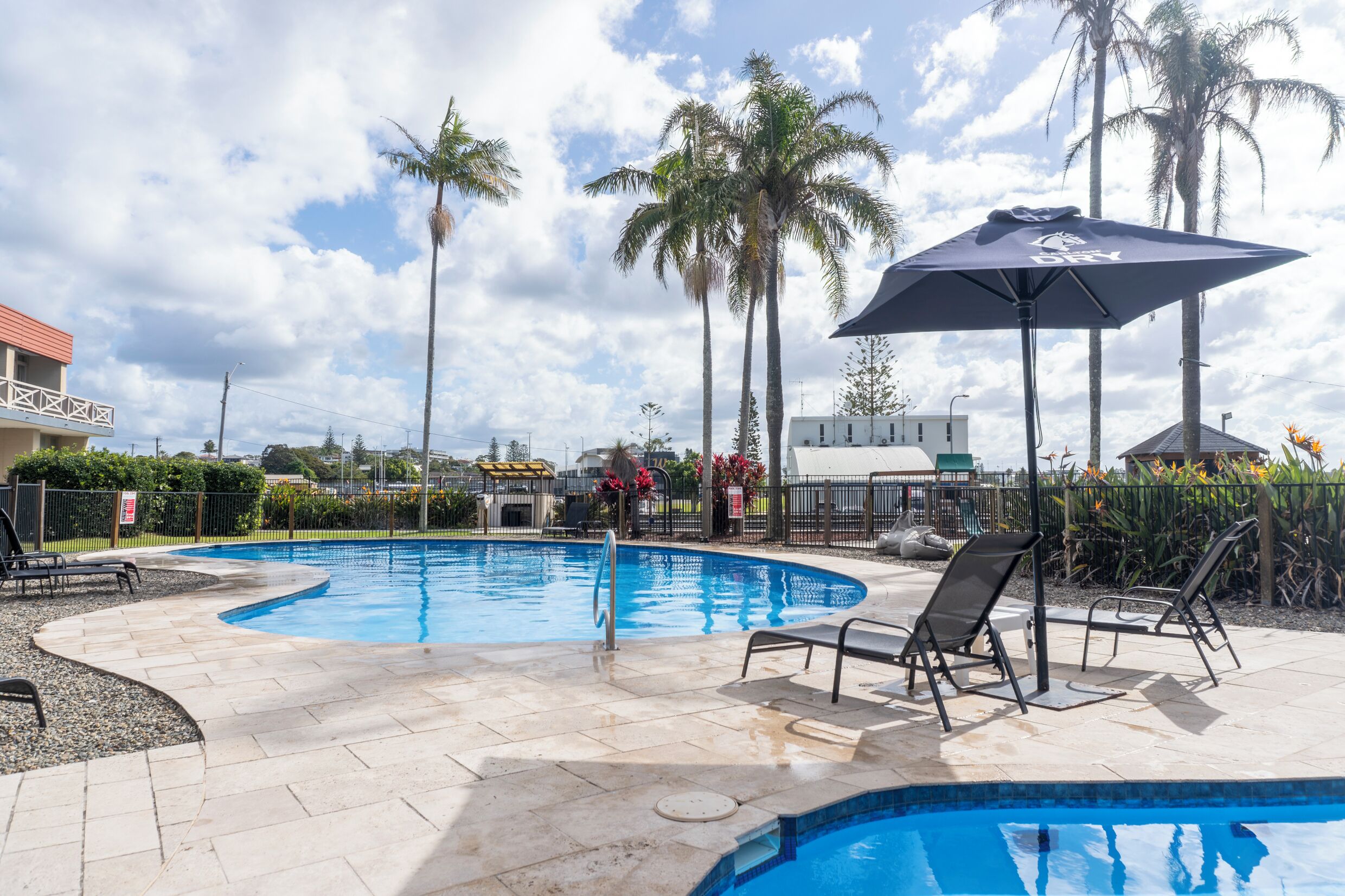 A swimming pool with lounge chairs and umbrellas, surrounded by palm trees and a fence.