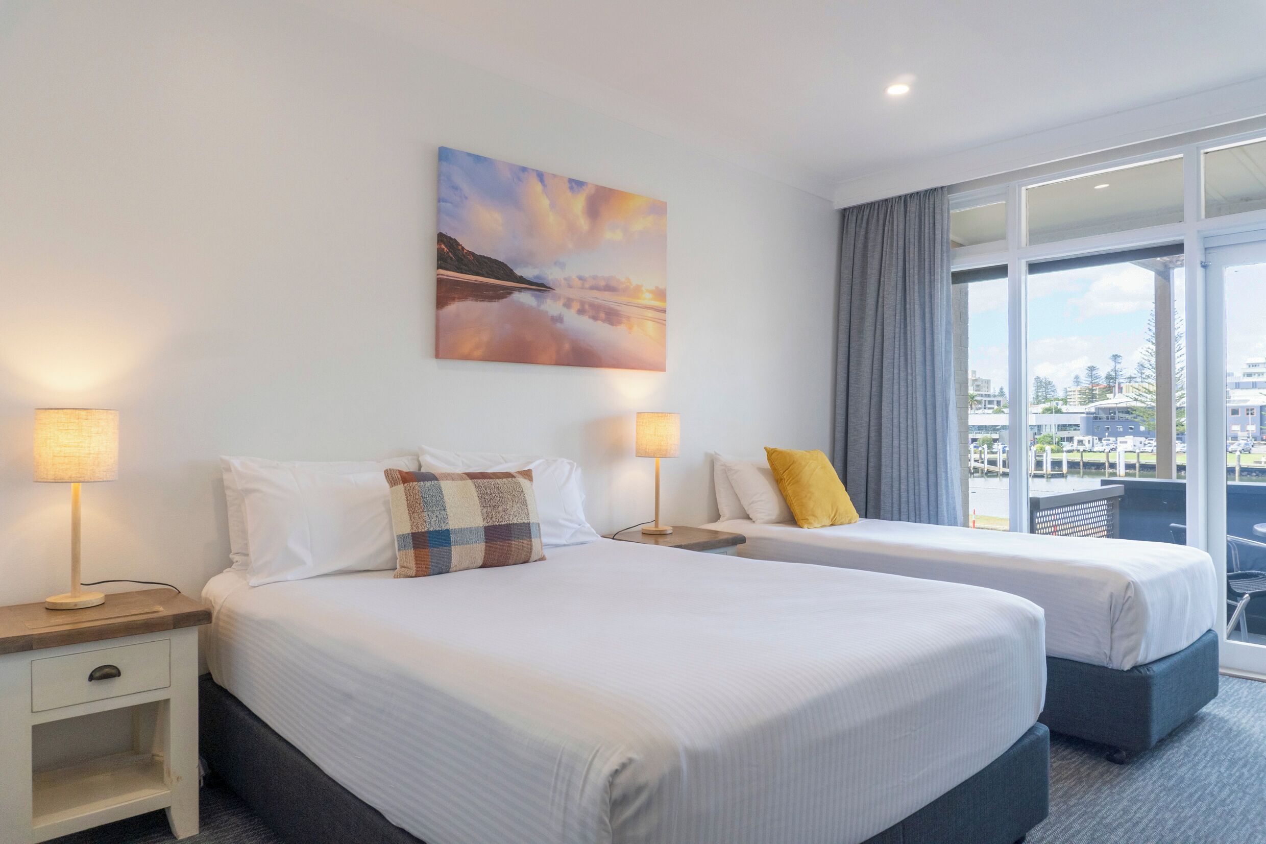 Ein Hotelzimmer mit zwei Betten, Blick auf die Stadt und einem Sonnenuntergangsgemälde an der Wand.