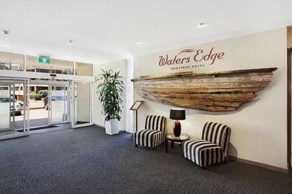 A reception area with a sign reading "Walters Edge Boutique Hotel," a large wooden wall decoration, two striped armchairs, a small table with a lamp, and a potted plant.