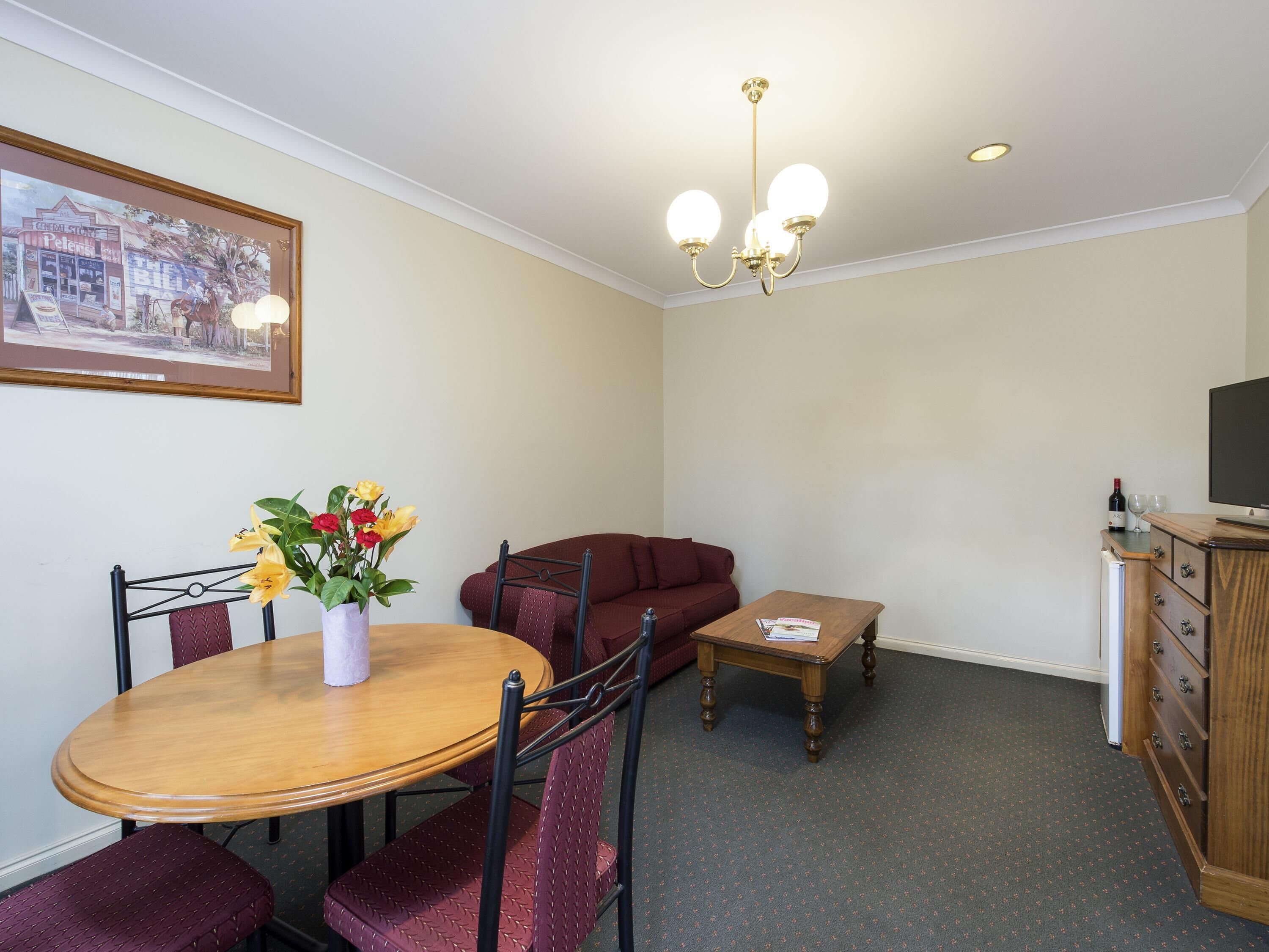 A dining area with a round table, chairs, a vase with flowers, a sofa, a small table with a book, a television on a cabinet, and a framed picture on the wall.