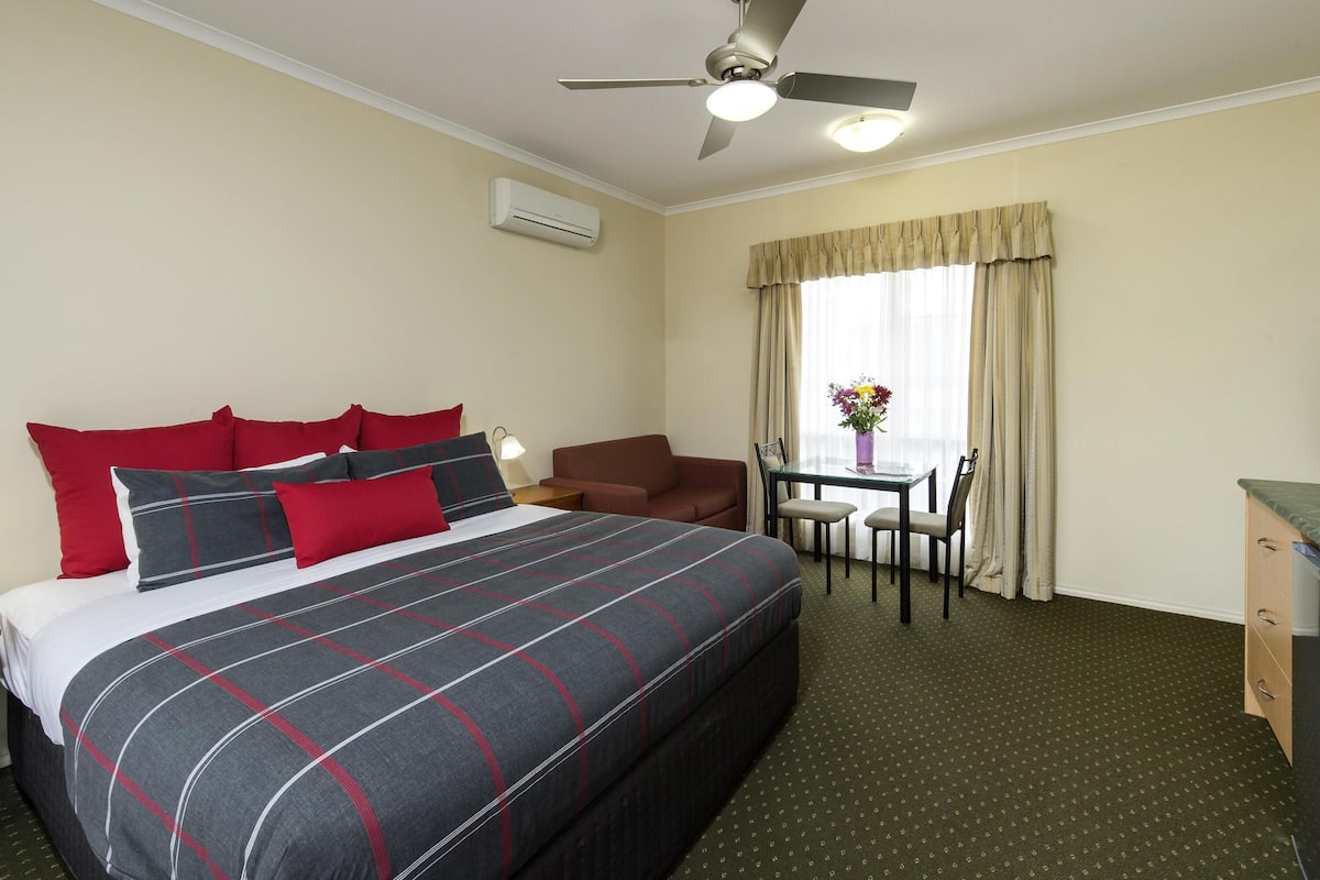 A hotel room with a bed, a ceiling fan, a table with chairs, and a window with curtains.