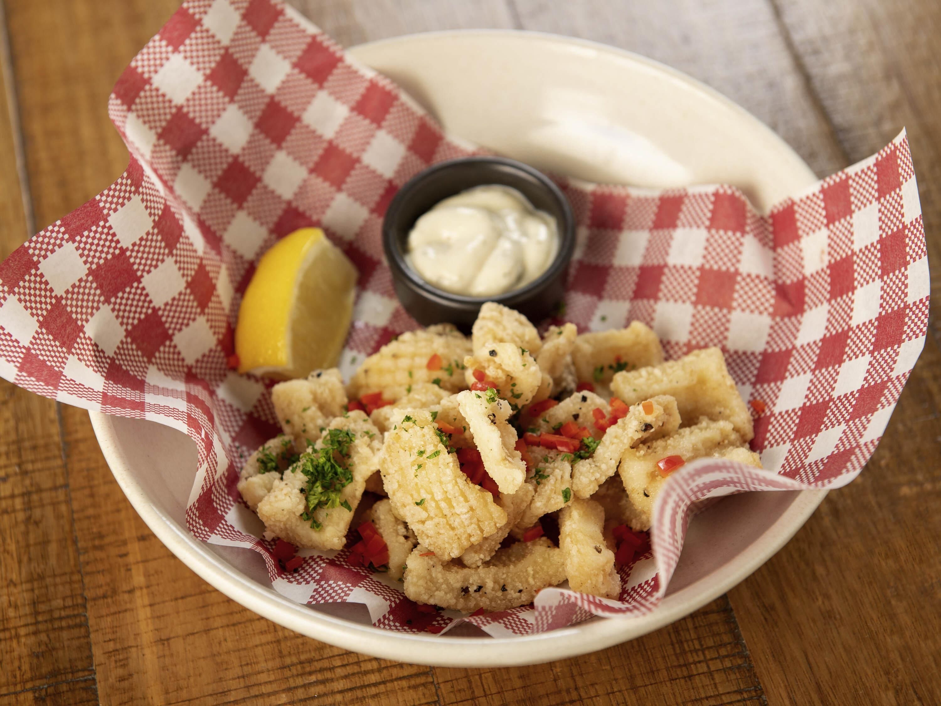 A bowl of fried cauliflower with a lemon wedge and a small bowl of sauce on a checkered paper.