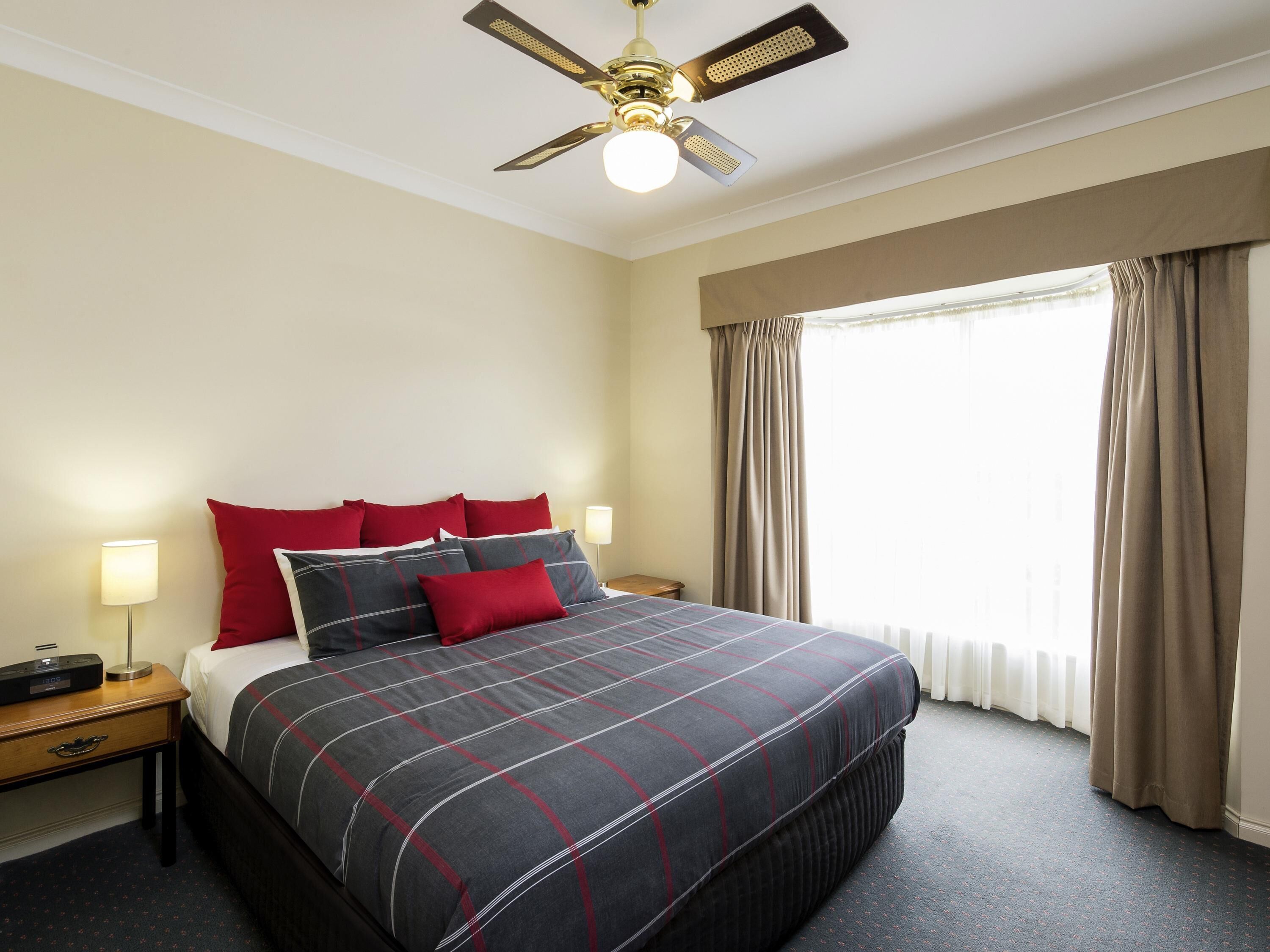 Une chambre d’hôtel avec un lit, une table de chevet, une lampe, un ventilateur de plafond et une fenêtre avec des rideaux.
