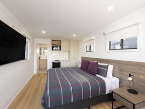 A modern hotel room with a large bed, a bedside table with a lamp, a built-in desk, a small kitchenette, and a TV mounted on the wall. - Breakfree Port Pirie (Solomontown)