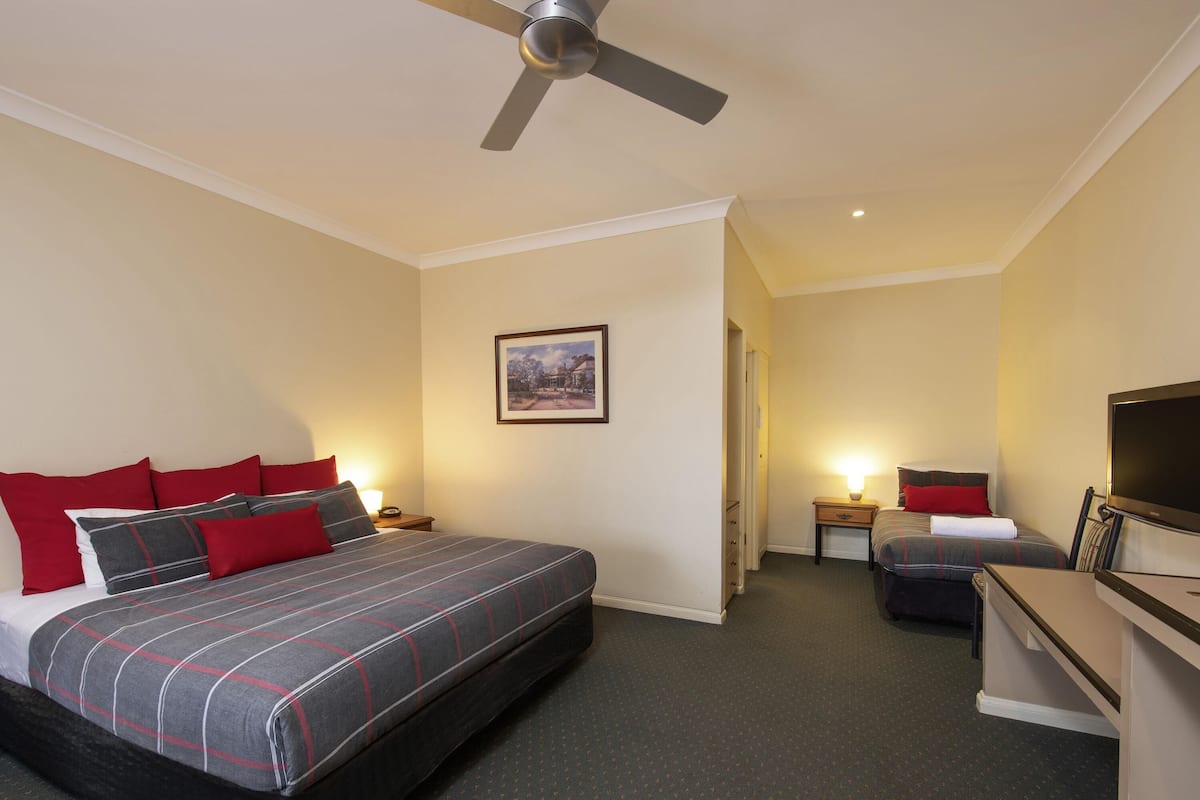 A hotel room with a large bed, a smaller bed, a desk with a television, a ceiling fan, and a framed picture on the wall.