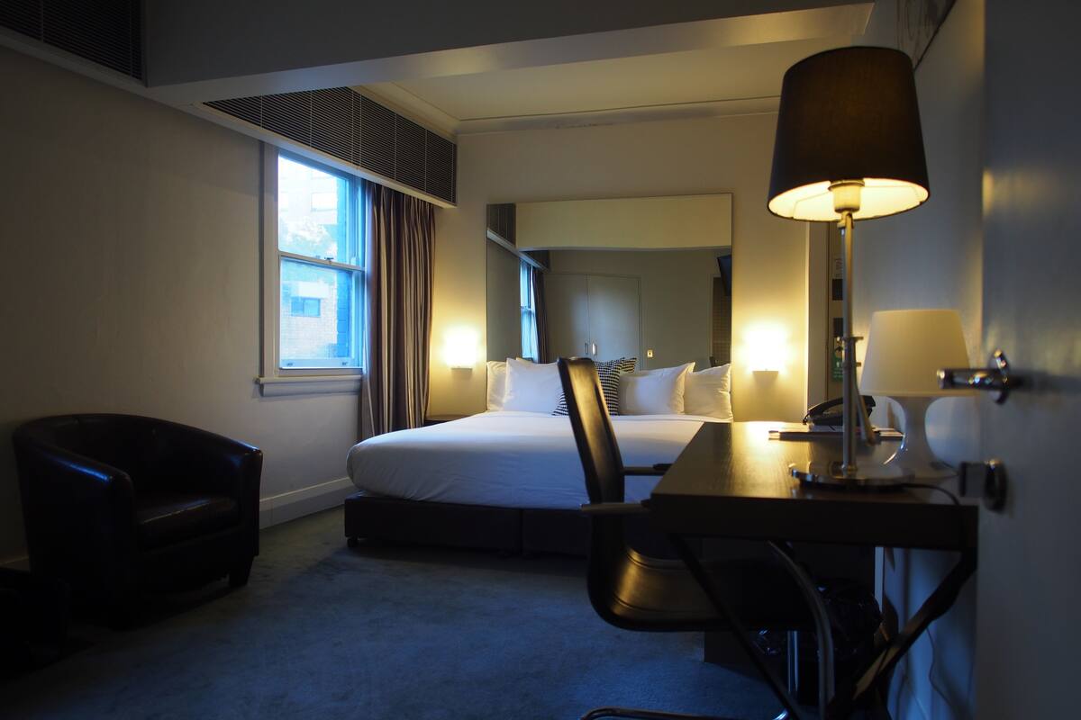 Habitación de hotel con cama, escritorio, silla, lámpara y ventana con cortinas.