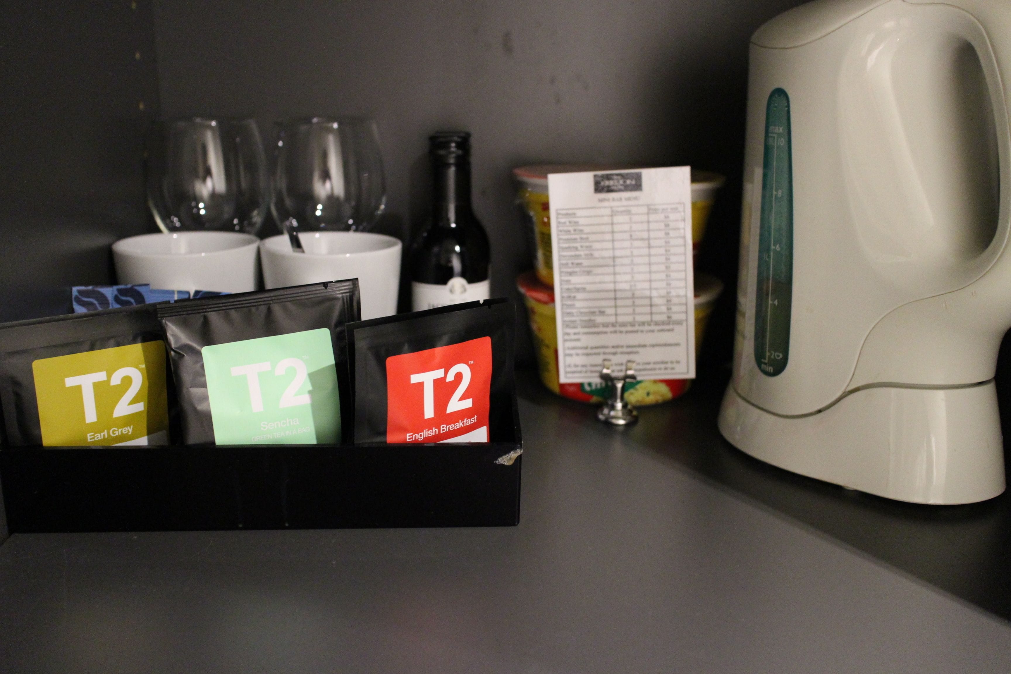 A kitchen counter with a coffee maker, three wine glasses, a wine bottle, and a T2 English Breakfast tea package.
