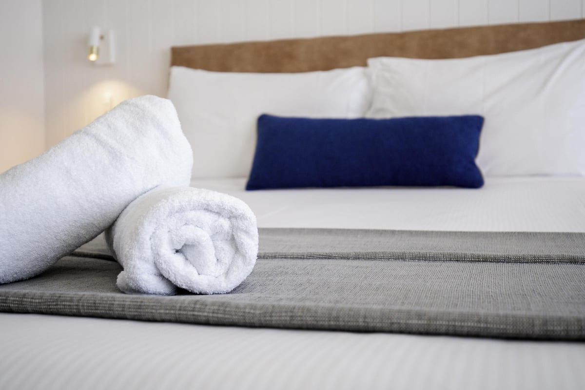 A neatly made bed with white and blue pillows, a rolled white towel, and a grey bedspread.
