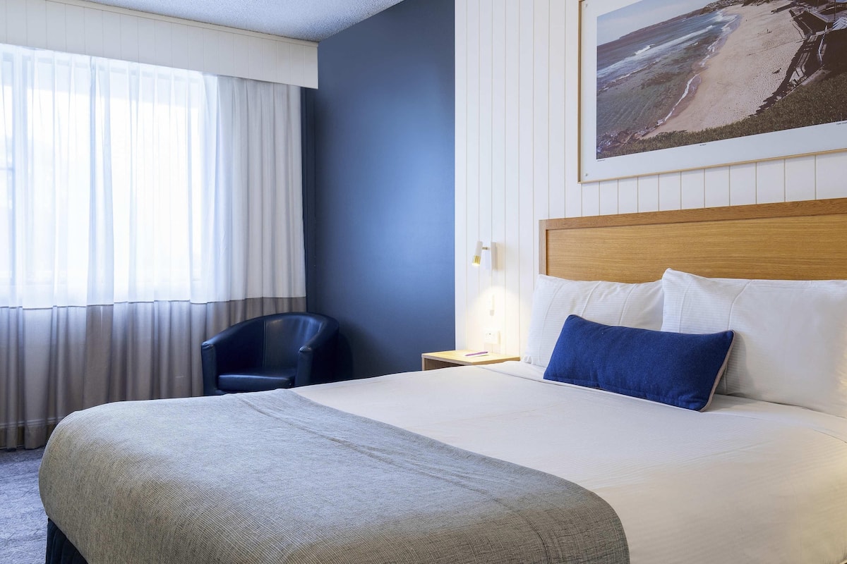 A hotel room with a bed, a chair, a nightstand, a lamp, and a painting on the wall.