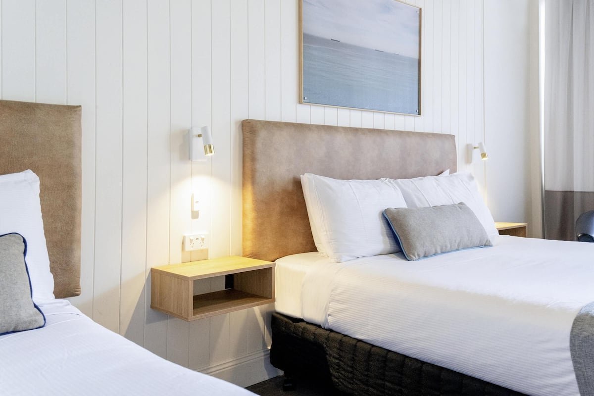 A hotel room with two beds, a wooden headboard, white bedding, a nightstand with a lamp, and a framed picture on the wall.