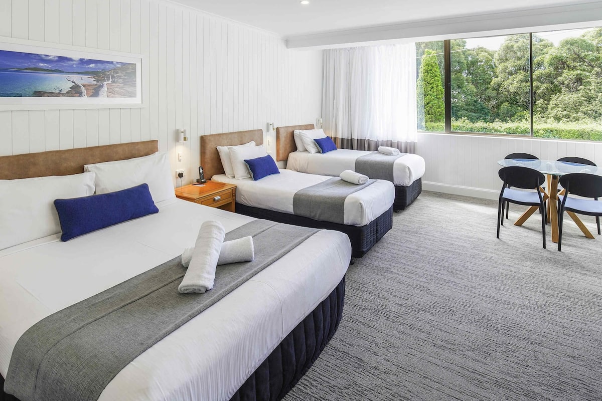 A hotel room with two beds, a dining table with chairs, and a large window with a view of greenery.
