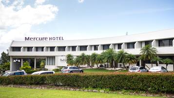 A modern hotel building with the name "Mercure HOTEL" on the facade. There are several parked cars in front, and palm trees are visible in the landscaped area.
